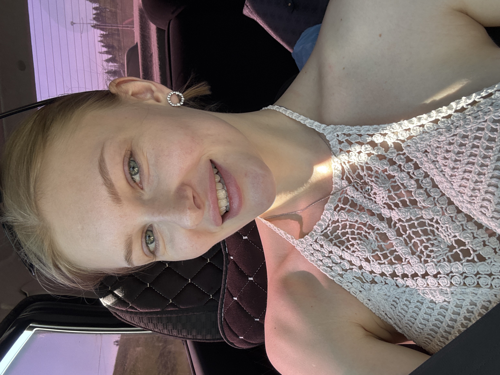 A fair-skinned woman with blonde hair pulled back is smiling while sitting in a car. She is wearing a white crocheted top and a small round earring. The interior of the car is dark, with a quilted leather car seat. The car window shows a blurred view of a landscape and a pink-tinted sky. There is no way to tell if the image was taken in Perm, Russia.