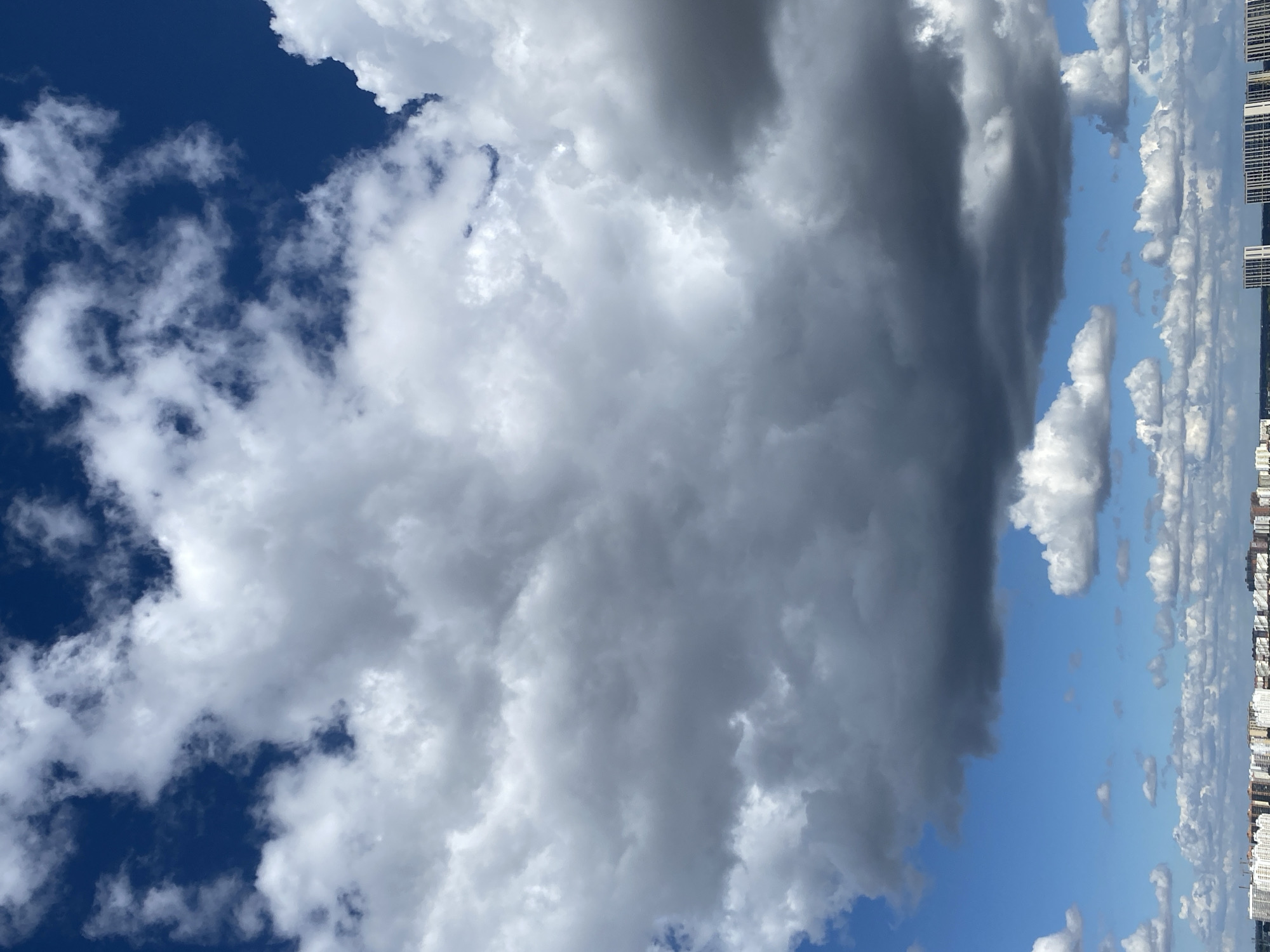 The image shows a sky filled with clouds above a city skyline. The clouds are varied in appearance, with some being dark and heavy, while others are white and fluffy. The sky itself is a deep blue. Below the clouds, a row of buildings stretches across the horizon, indicating an urban environment. The architecture suggests apartment buildings, characteristic of a modern city. The lighting indicates it is daytime, likely with the sun obscured by some of the denser cloud cover. The scene is static and devoid of any visible human activity or movement. Given the presence of a distinct style of apartment blocks visible in the foreground and common in Russia, this scene is likely located in the city of Krasnodar, Russia.