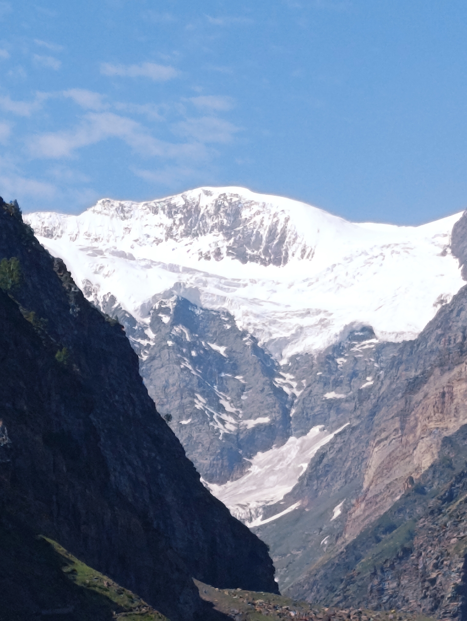This image captures a majestic mountain landscape with a snow-capped peak dominating the background. The mountain face is rugged, with visible rock formations and patches of snow. In the foreground, dark, steep cliffs flank a valley leading towards the snowy mountains. Hints of green vegetation can be seen on the lower slopes of the cliffs, suggesting a mountainous region with varying altitudes. The sky above is a clear blue with scattered white clouds. The scene appears to be a natural, remote location, likely a part of the Indian Himalayas.