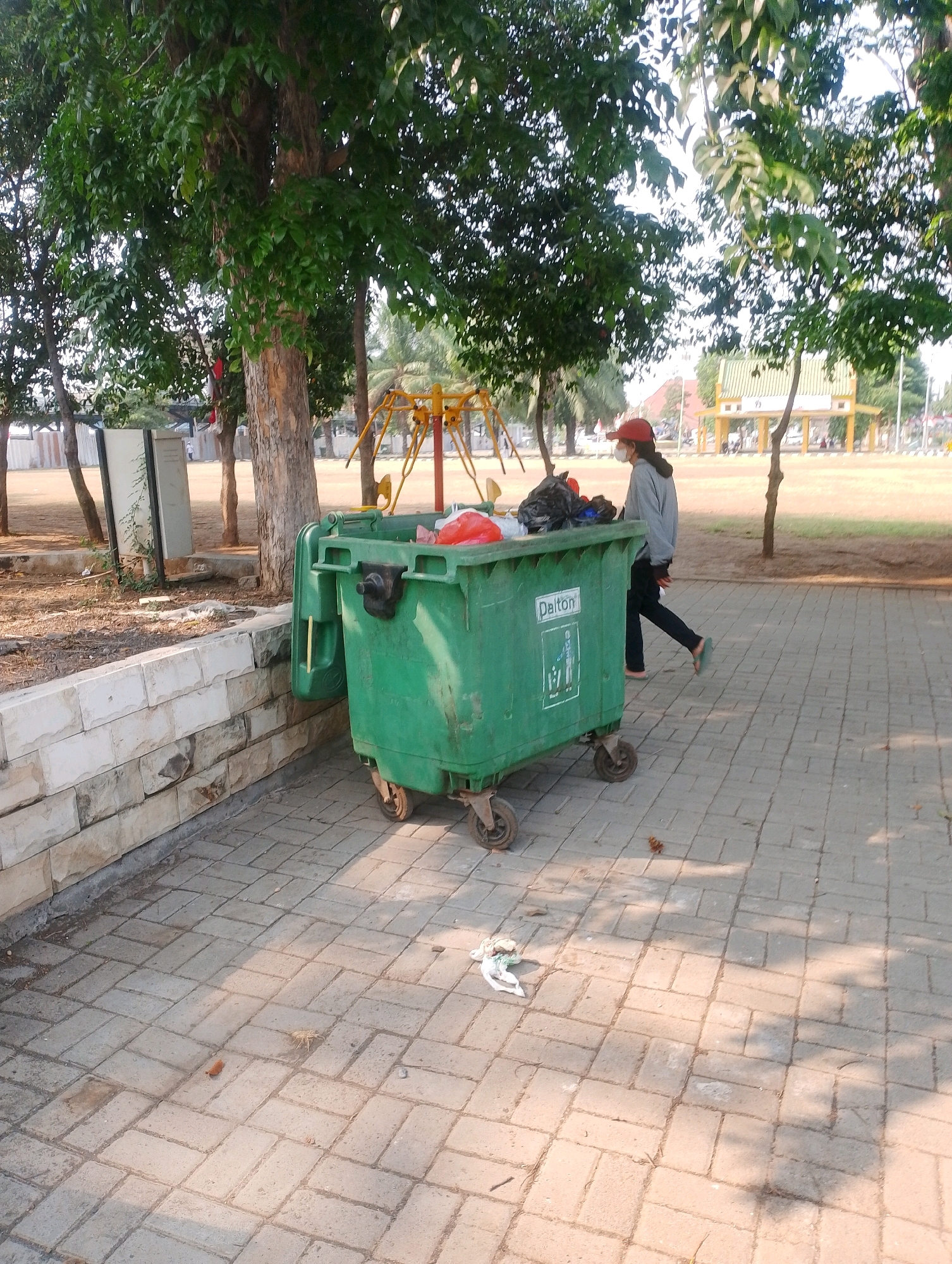 The image shows a green, wheeled dumpster full of trash, positioned on a paved walkway in a park in Probolinggo, Indonesia. A person wearing a red cap, grey hoodie, dark pants, and sandals walks away from the dumpster towards the right side of the frame. They appear to be wearing a white face mask. The dumpster has the brand name "Dalton" visible on its side. In the background, there are trees, a playground with yellow swings, and a yellow-roofed building with white pillars. The ground is paved with interlocking bricks, and there is a low stone wall to the left of the dumpster. Sunlight casts shadows across the paved area.