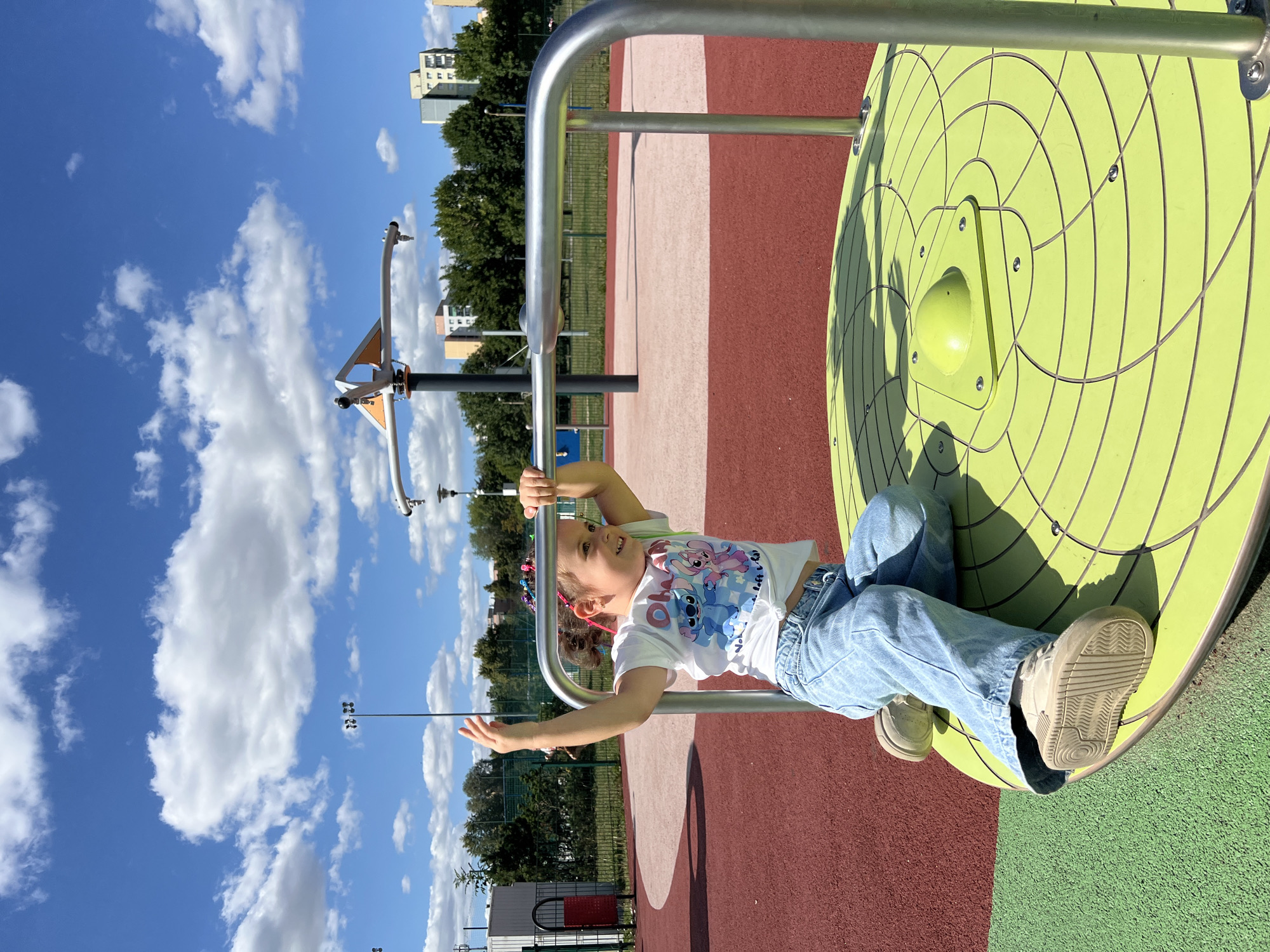 A young girl with braided hair, wearing a white t-shirt with cartoon characters and light blue jeans, is sitting on a bright green merry-go-round in a playground. She is holding onto the metal railing and looking to her right with a smile. The playground has red and pink rubber safety surfaces. In the background, there are trees, apartment buildings, and a blue sky with white clouds. The scene appears to be located in the city of Lublin, Poland, as indicated by the architectural style of the buildings.