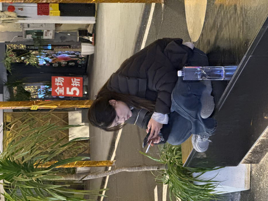 A young East Asian female sits in a semi-squat position on a dark, reflective urban bench or ledge in Chongqing High-tech Zone, China, under artificial evening illumination. She wears a black puffer jacket, a grey long-sleeved shirt, blue wide-leg jeans, and grey athletic shoes. Her dark hair is pulled back. She is focused on a black smartphone held in both hands. An unlit cigarette is held between the index and middle fingers of her left hand. To her right, on the same reflective surface, stands a clear plastic water bottle with a white cap. The background features a street or alleyway. To her immediate left is a tall potted plant with long green leaves. Further back, two gold-wrapped pillars frame a storefront entrance. A prominent red sign with white Chinese characters "全场 5折" (50% off storewide) is visible between the pillars. Beyond, storefront interiors display various illuminated items and additional potted greenery. The ground is a dark paved surface.