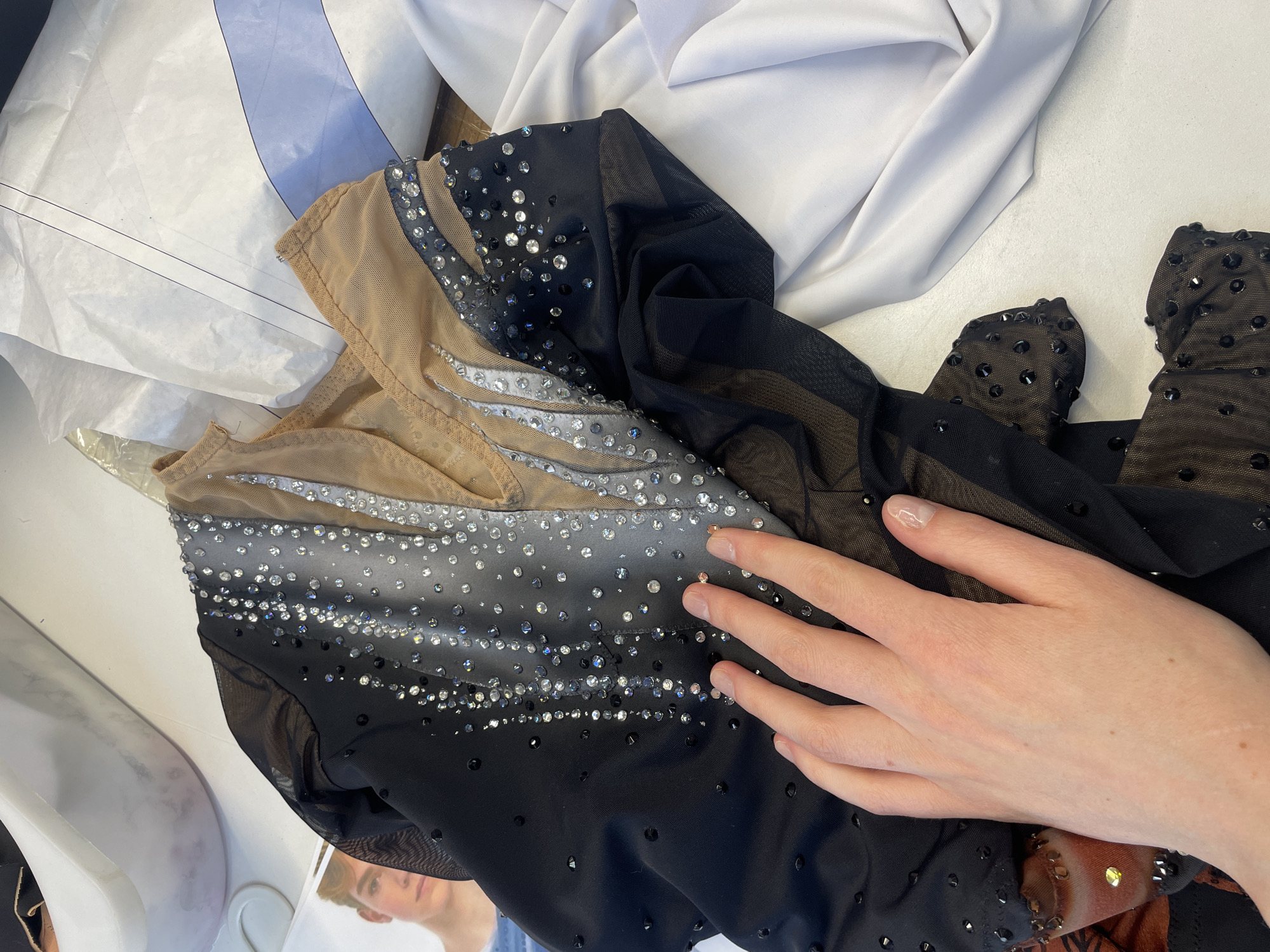 A hand rests on a black and tan performance costume adorned with rhinestones. The costume is laid out on a white surface, possibly a table, near some white fabric and what appears to be sewing patterns. The background shows a blurred white bin. The image is taken from a slightly elevated angle, focusing on the costume and the hand. There is no indication of a location in Saint Petersburg, Russia, as the image provides no discernible landmarks or contextual clues for such a determination.