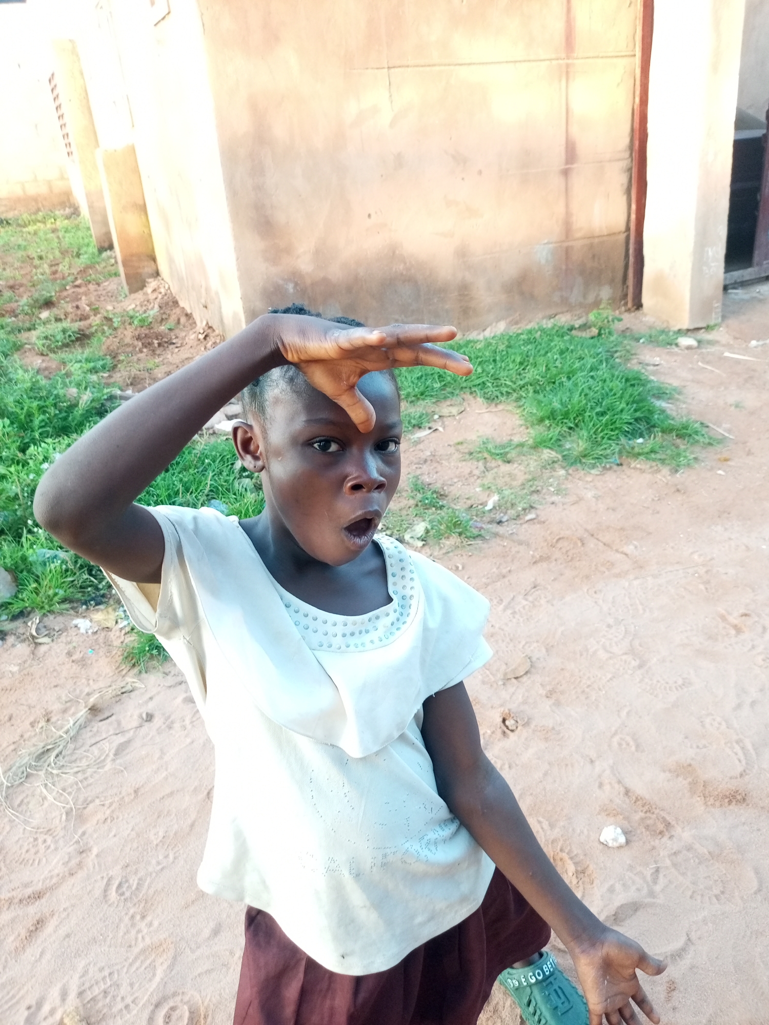 In a dusty, sandy outdoor area in Tempure, Nigeria, a young Black girl is pictured. She is wearing a light-colored, short-sleeved top with a decorative neckline and a dark skirt. Her right arm is raised, with her hand shielding her eyes as if looking into the distance. Her mouth is open in a surprised or exclamatory expression. In the background, a rough, beige-colored building stands, with patches of green grass and some debris on the ground. The overall impression is one of candid observation in a simple, rural or semi-urban setting.