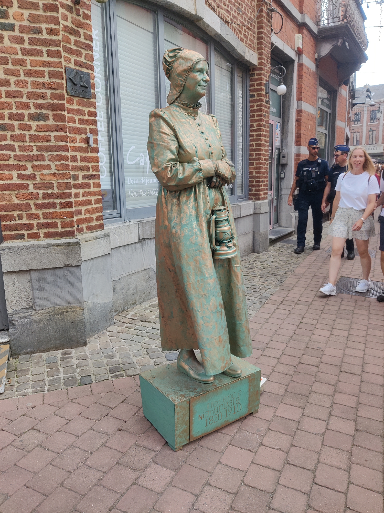 A statue of a woman dressed in historical attire, painted to resemble bronze, stands on a brick sidewalk in Marche-en-Famenne, Belgium. The statue is dressed in a long dress and a bonnet, and holds a lantern in its right hand. The base of the statue is a teal box with "Florence Nightingale 1820-1910" inscribed on it. In the background, two police officers are walking, and a woman is smiling as she walks towards the camera. A brick building with a window and signage for "COFFEE CORNER" is visible behind the statue. The sidewalk is paved with red bricks, and there are cobblestones in the background.