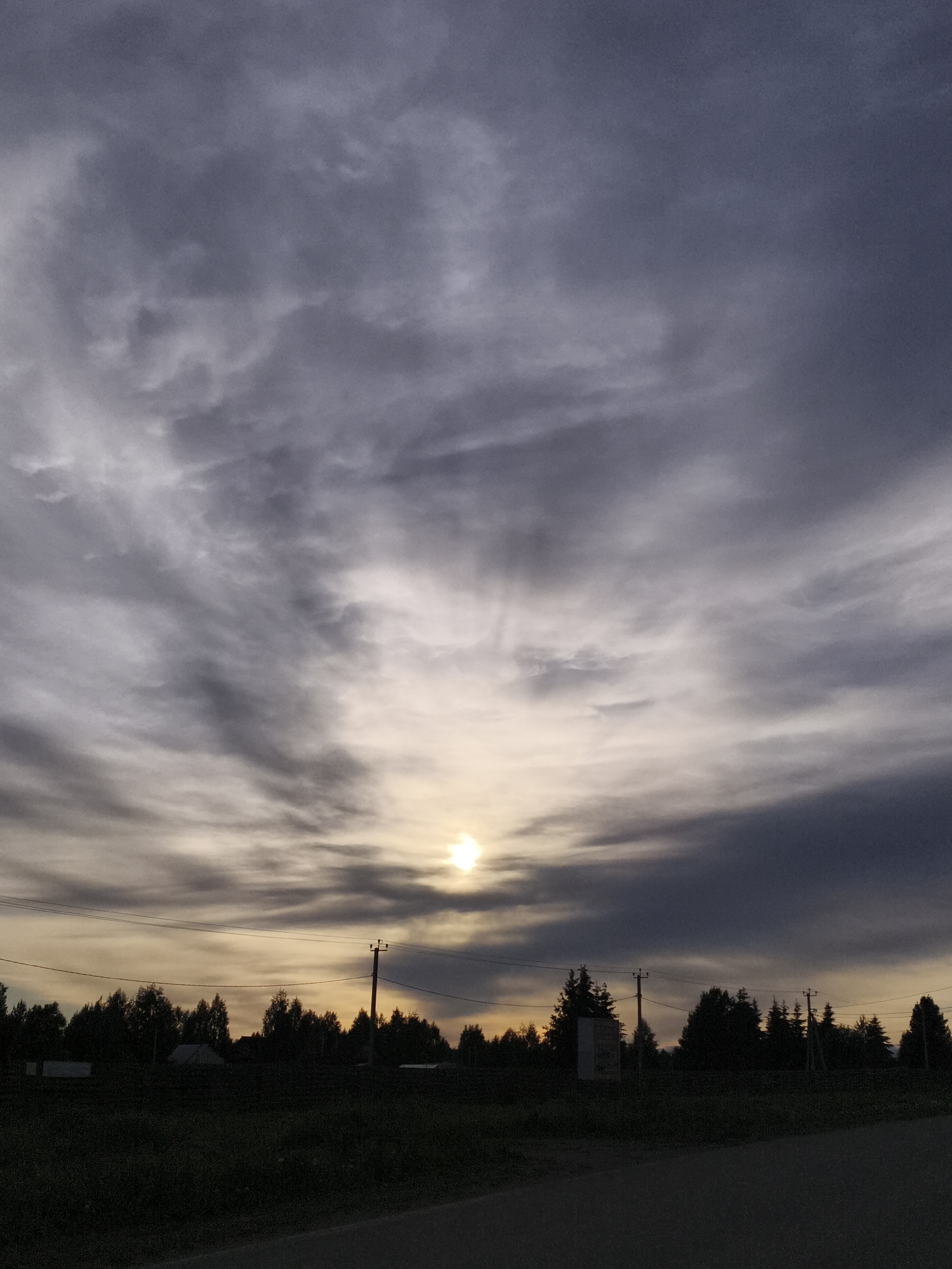 A cloudy sky dominates the view, with the sun partially obscured by the clouds near the horizon. A line of trees and some buildings form a silhouette against the brighter sky. Utility poles with wires run across the middle ground of the image. The foreground consists of a patch of grass and a paved road. The picture may be taken in the general vicinity of the city of Русский Вожой, Russia, due to the types of buildings and landscapes.
