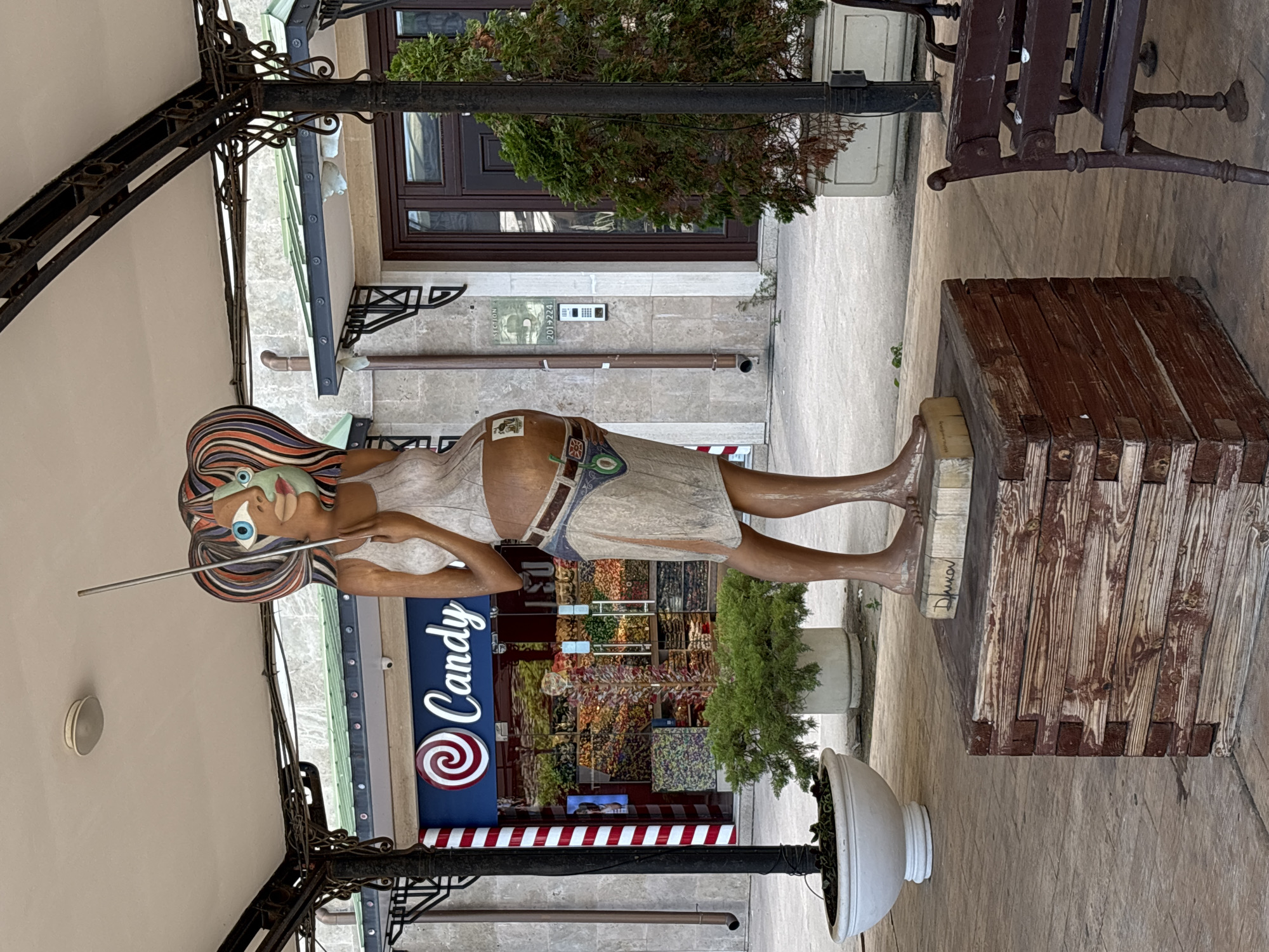 This is a vertical, full-shot photograph of a whimsical sculpture of a woman with vibrant, multi-colored hair and an exaggerated, cartoonish face. She is dressed in a simple light-colored dress and appears to be holding a long, thin object, possibly a straw or a wand, to her lips. The sculpture is standing on a wooden platform.
In the background, there is a shop with a large sign that reads "Candy" in white lettering with a red and white swirl logo. The shop window displays an abundance of colorful candies. Adjacent to the shop, a green bush or small tree is planted in a white pot.
To the right of the sculpture, a dark wooden bench with intricate metalwork is visible, suggesting an outdoor seating area. The ground is paved with textured stones. A dark metal awning structure extends over the scene from the left. The overall setting appears to be a pedestrian walkway or an outdoor commercial area. The lighting suggests it is daytime, likely overcast or shaded due to the awning. The presence of the candy shop and the playful sculpture creates a cheerful and inviting atmosphere. The sculpture has some text on its base, which is difficult to read clearly but appears to be a name or signature. There is also a sign on the building in the background with some text and numbers that are too small to discern.