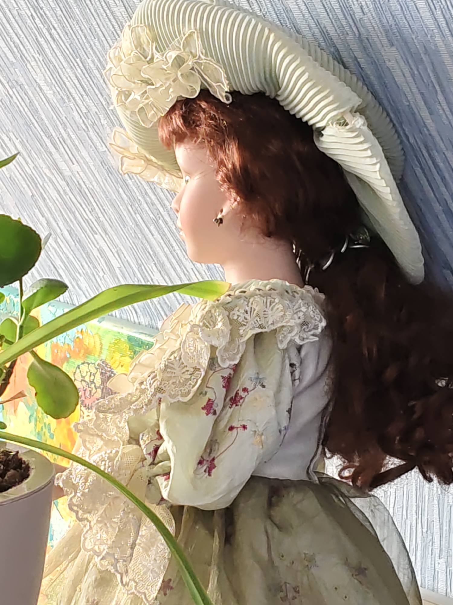 This is a close-up, eye-level, slightly upward-angled, medium shot of a porcelain doll with long, auburn hair. The doll is wearing a voluminous light green, pleated bonnet with ruffled lace trim, and a matching light green dress with floral embroidery and ornate lace detailing on the collar and sleeves. A portion of a green potted plant with broad leaves is visible in the foreground on the left. The background consists of a textured, pale blue surface with vertical lines, and a blurred colorful artwork or fabric behind the doll. The scene appears to be indoors, possibly near a window, with natural light illuminating the doll and plant. There is no indication of the city of Rostov-on-Don, Russia, in the image.
