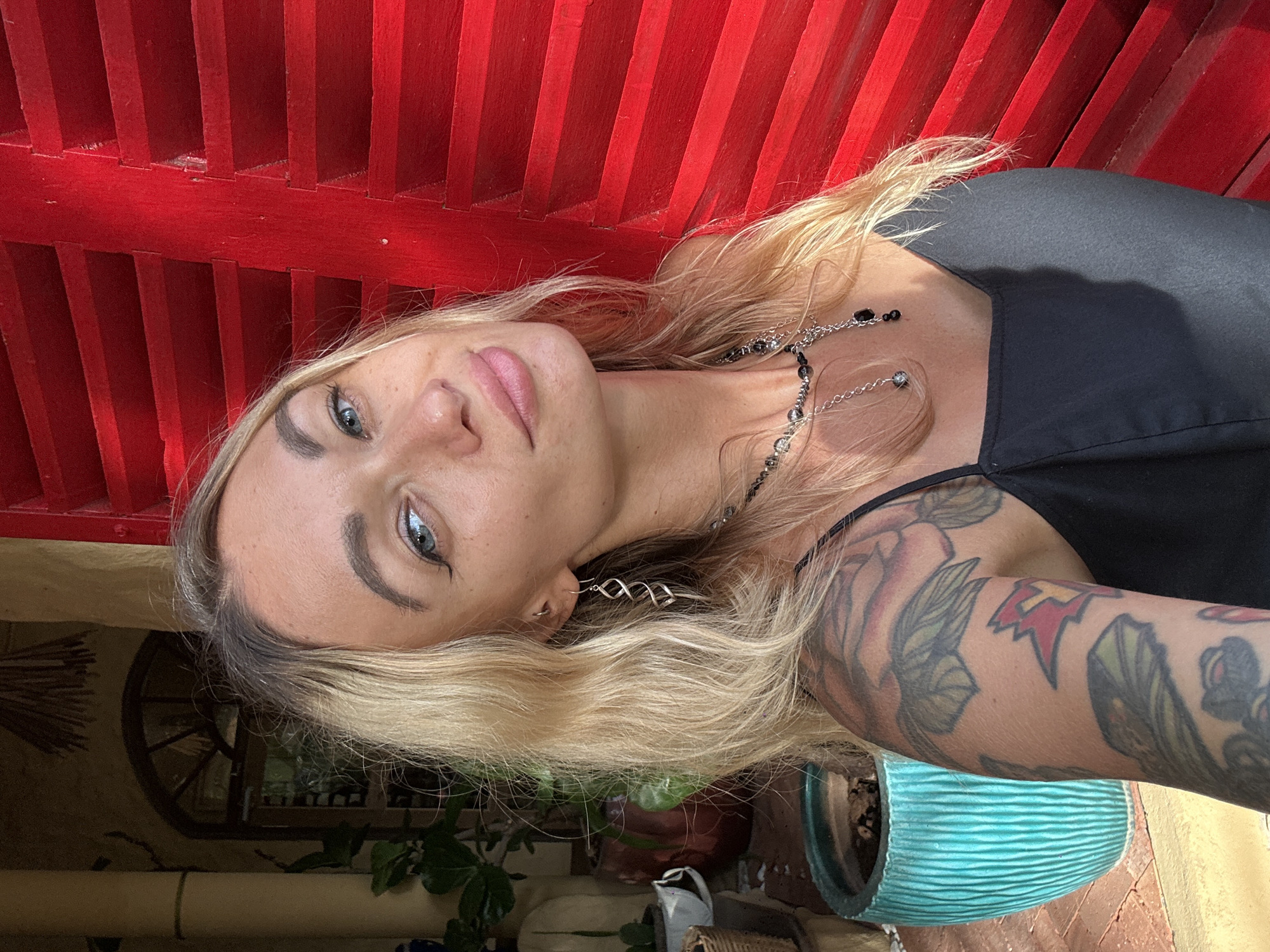 In Mũi Né, Vietnam, a woman with blonde hair and blue eyes is captured in a selfie. She has freckles across her nose and cheeks, and is wearing a black tank top, a silver necklace, and a silver spiral earring. Her left arm is covered in tattoos. She stands in front of vibrant red shutters, with a glimpse of a pillar, an arched window, potted plants, and a turquoise planter visible in the background. The scene appears to be outdoors, possibly on a patio or verandah, bathed in natural light.
