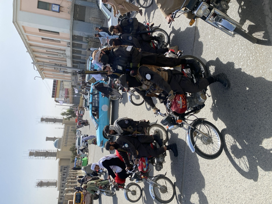 The image depicts a busy daylight street scene in Kandahar, Afghanistan. In the mid-background, the Shrine of the Cloak (Kherqa Sharif) Mosque is prominent, a large beige structure with a blue dome and four minarets, two of which are under construction and covered in scaffolding. To the right of the mosque, a multi-story building with a peach-colored facade and columns is visible.
The immediate foreground and mid-ground are densely populated with men on motorcycles and other vehicles. A man wearing dark tactical clothing, a tactical vest, and a dark face mask stands on the seat of a red motorcycle on the right side of the frame, holding an AK-style assault rifle vertically in his left hand. To his left, another man in similar tactical gear and a tactical vest rides a red motorcycle. Further left, two men occupy another red motorcycle; the driver wears dark clothing and a cap, while the passenger, dressed in dark clothing and a dark headscarf, carries an AK-style rifle slung across his back. A man with a distinctive white and brown patterned headscarf and a white headband around his forehead rides a red motorcycle to the left. Multiple other individuals on motorcycles are present, many armed with AK-style rifles and wearing a mix of dark tactical uniforms and traditional Afghan attire.
The street itself is paved asphalt, displaying strong shadows consistent with bright sunlight. Traffic includes numerous motorcycles (predominantly red Honda models), a light blue sedan with a white circular logo on its side, other cars, and pedestrians, indicating a bustling urban setting with a visible presence of armed individuals.