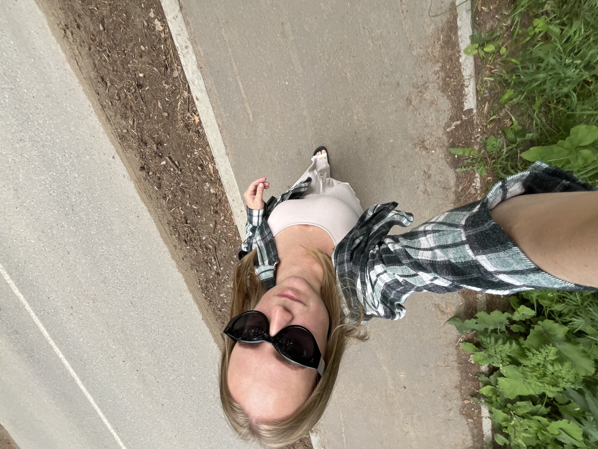 A woman is taking a selfie on a paved walkway next to a road in Chaikovsky, Russia. She is wearing black sunglasses, a light-colored top, and an open green and black plaid shirt. Her left arm is extended, holding the camera. The walkway is bordered by a dirt and debris area, and on the other side by a road with a white line marking. Green vegetation is visible along the edge of the walkway.