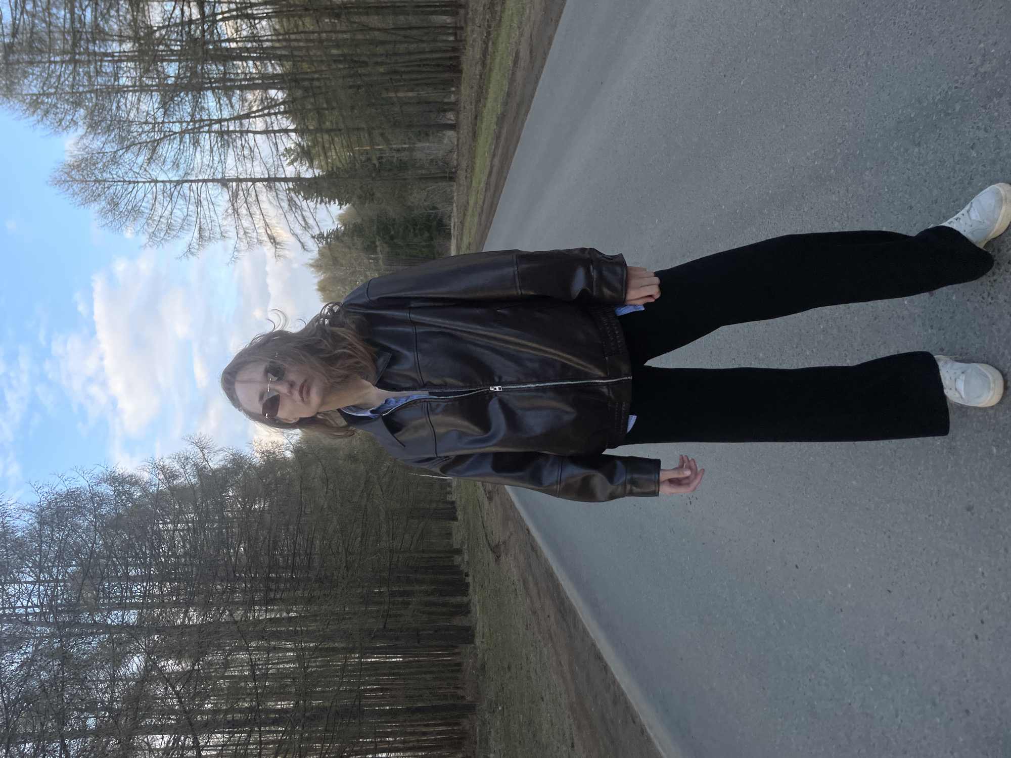 A full-length photograph captures a person standing on a grey asphalt road in a natural, possibly rural setting. The subject, likely female, has long, wavy brown hair and is wearing dark sunglasses, looking directly towards the camera. They are dressed in a dark brown or black leather-look jacket, black wide-leg trousers, and light-colored sneakers, adopting a casual stance.
The scene depicts a straight, grey asphalt road stretching into the distance. On both sides of the road, dense lines of trees with bare branches are visible, suggesting either late autumn, winter, or early spring. The sky above is bright blue with scattered white clouds. The overall impression is one of open space and natural surroundings, possibly a forest road. The time of day appears to be daytime with clear weather. Given the provided context, the location is Podshivalovo, Russia. There is no visible text within the image.