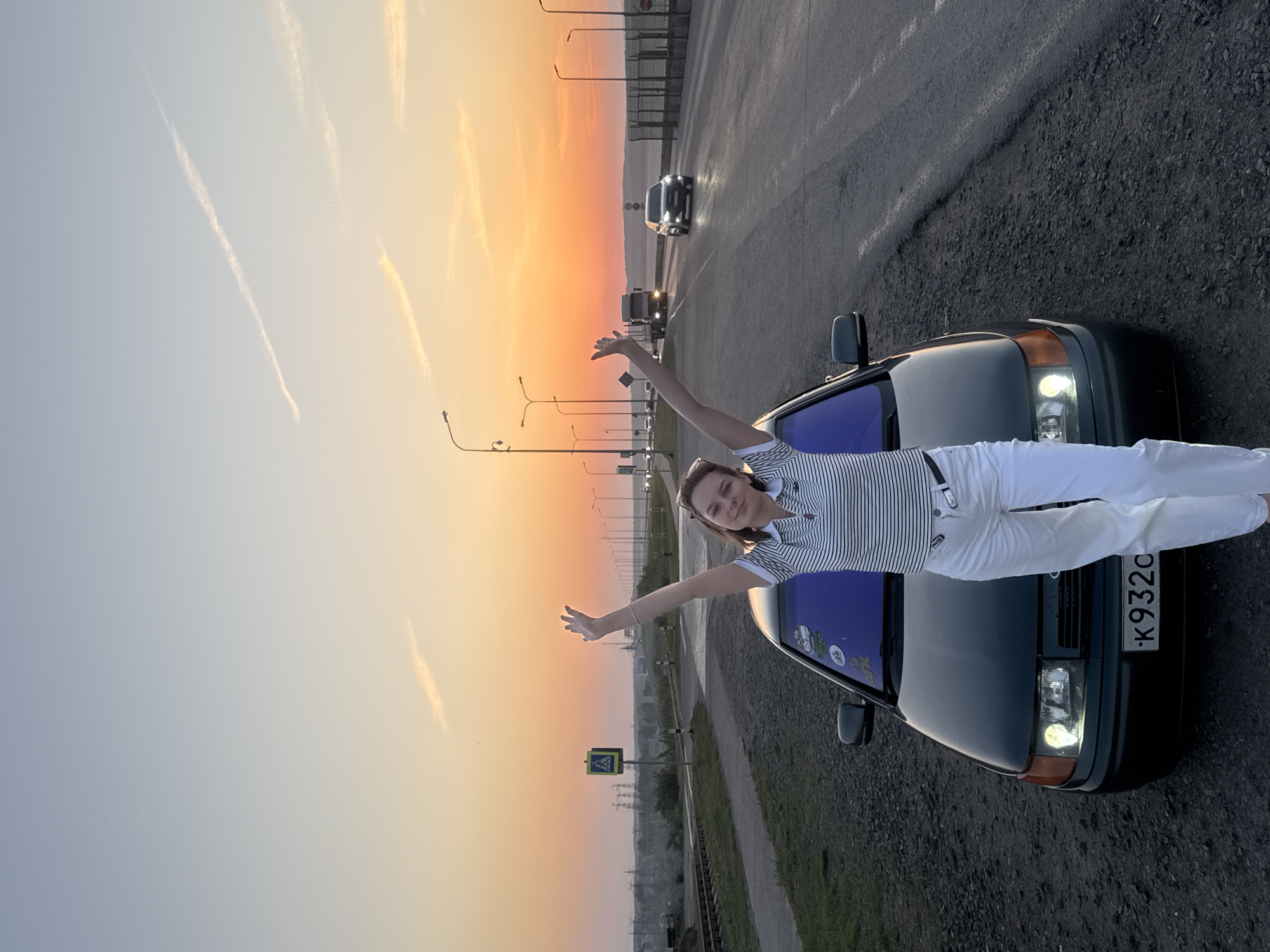 A young woman stands on the hood of a dark-colored car, extending her arms, in Chaikovsky, Russia. She wears white pants and a striped shirt. The car faces a road with cars and streetlights leading towards a vibrant sunset. To the side of the road are train tracks and a pedestrian crossing sign. The car's license plate reads "K932CO." The sky blends from pale blue to orange near the horizon, with contrails visible. The road surface appears to be asphalt and gravel.