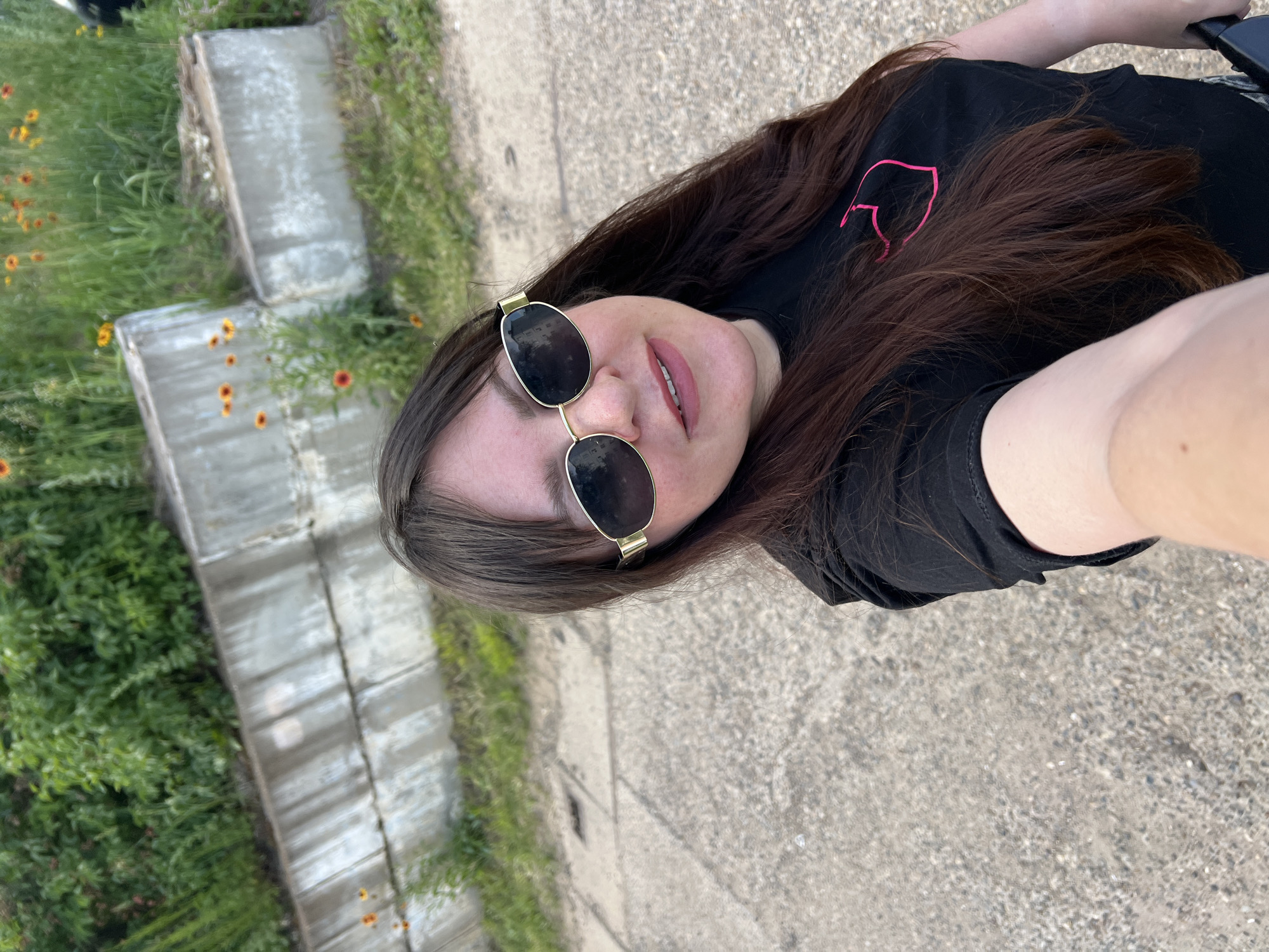 A woman is taking a selfie outdoors. She is wearing a black t-shirt with a pink outline of a heart on the left side of her chest and black shorts. She also has on large, round, dark sunglasses with gold frames. The woman has long, dark brown hair that is spread across her shoulders and down her back. Her mouth is slightly open, and she is looking up at the camera. The background shows a paved surface with patches of green grass and some small, orange and yellow flowers. There are also some weathered concrete structures in the background. The lighting suggests it is daytime.
