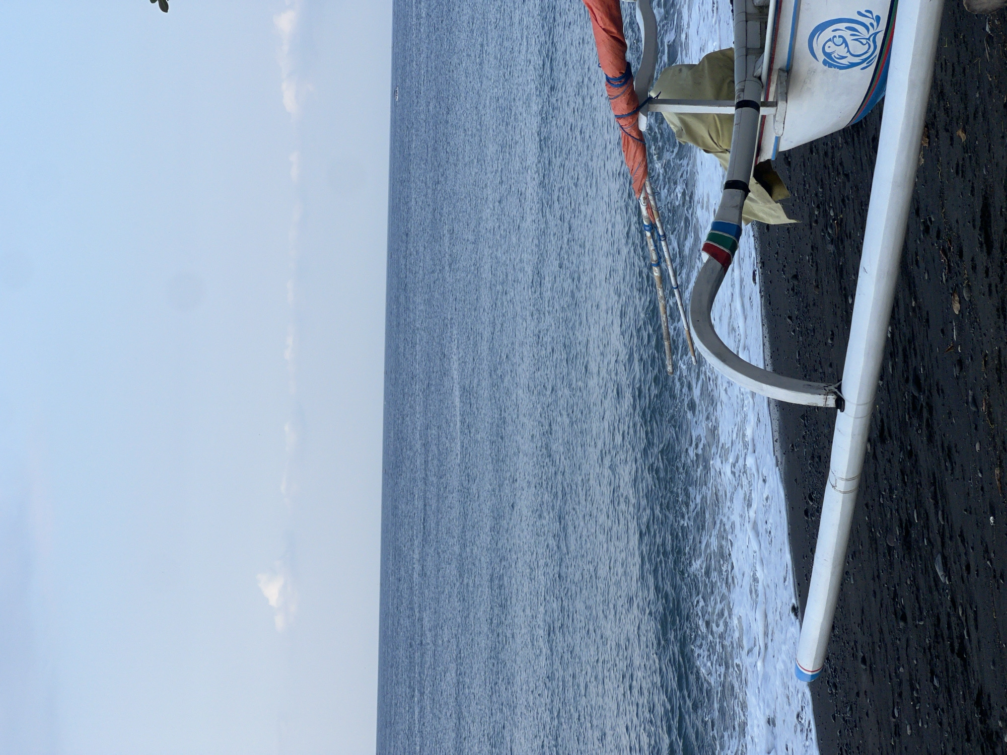A low-angle, medium shot captures a traditional Balinese fishing boat partially beached on a dark, pebble-strewn shore. The boat, predominantly white with blue trim and a stylized blue dolphin decal, is positioned to the right of the frame, facing towards the left. Its outriggers extend horizontally from the hull, with one end prominently visible in the foreground. A rolled-up orange tarp is secured to the outrigger structure with blue rope. The water, a deep blue, stretches to the horizon under a pale blue sky dotted with faint white clouds. Gentle waves with white foam lap at the shore. The scene is devoid of people. This location is consistent with coastal areas in Abang, Indonesia, known for its fishing communities and traditional boat building.