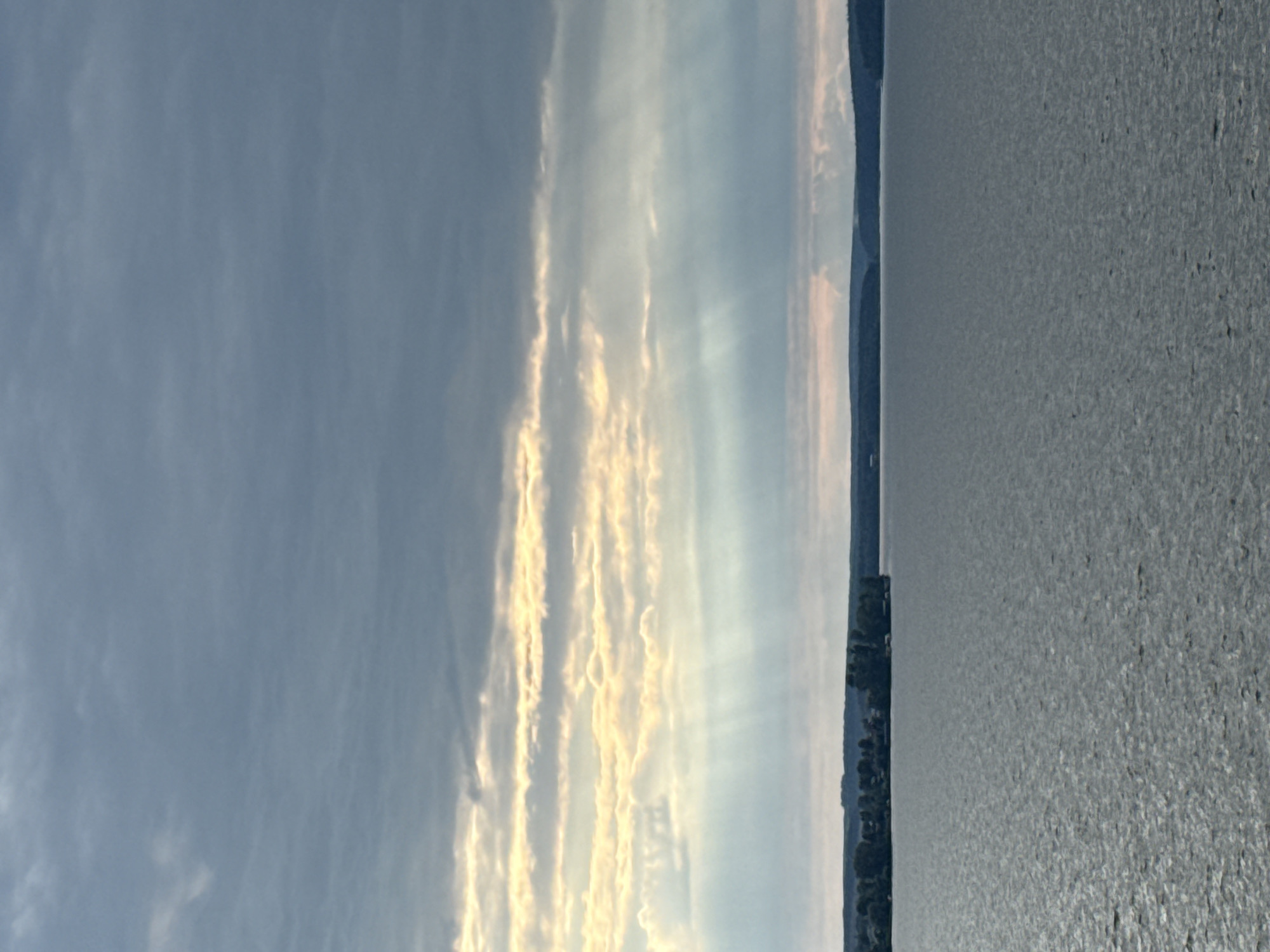 The image depicts a wide expanse of water, likely a lake or a large river, with a tree-lined shore visible in the distance. The sky is filled with dramatic clouds, with rays of sunlight piercing through in places. The water is choppy, with a textured surface that reflects the light. The overall scene suggests a natural landscape, possibly a scenic view from a boat or a vantage point overlooking the water. The location in Votkinsk, Russia, is not explicitly identifiable from the visual elements alone, but the description suggests a setting consistent with the natural environment of the region.