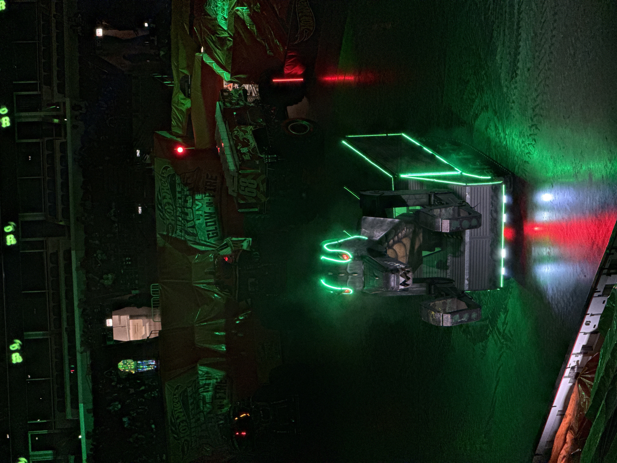 This is a low-light, high-angle exterior shot of a monster truck event. The scene is dominated by dark colors, with bright green and red neon lights illuminating parts of the arena floor and several vehicles. In the foreground, a monster truck designed to look like a dinosaur is positioned center frame. It has neon green lights outlining its head and body. The dinosaur truck is resting on a platform with metallic detailing. To the right and slightly behind the dinosaur truck, another monster truck, numbered "68," is visible. This truck has a darker design with red accents and is partially obscured by a banner that reads "GLOW-N-FIRE" and "HOT WHEELS MONSTER TRUCKS LIVE." In the background, bleachers filled with an audience are dimly lit, and a person wearing a brightly colored, illuminated costume stands near an exit sign labeled "EXIT 110." The overall environment suggests an indoor arena, possibly the United Center in Chicago, United States of America, given the context of large-scale entertainment events often held in such venues. The floor of the arena appears to be dark and possibly covered in dirt or a similar material.