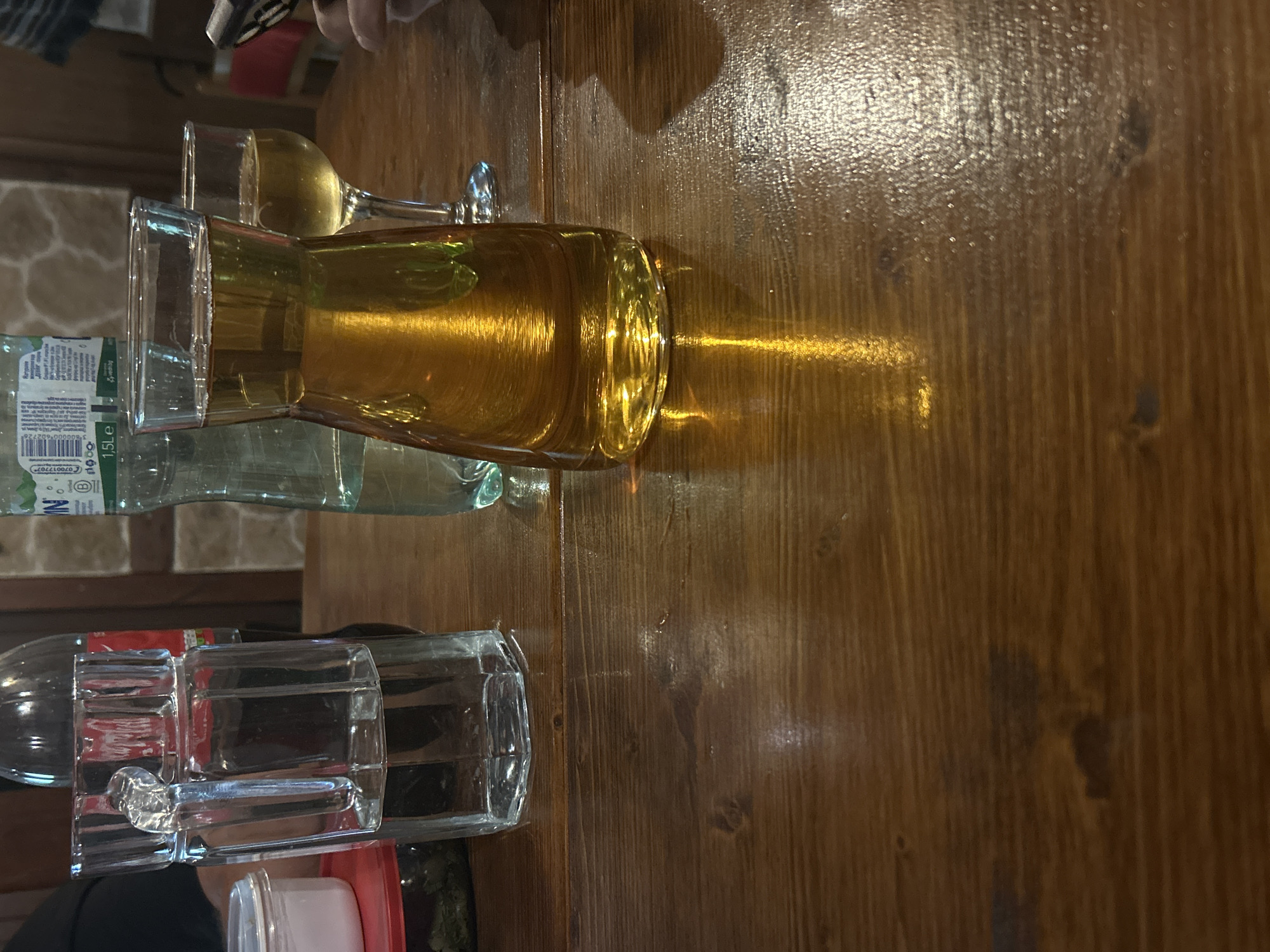 The image shows a table at an establishment in Mirovyane, Bulgaria. Several glass containers are present, including a carafe of an amber-colored liquid (likely a beverage) and a water bottle. There are also drinking glasses and what appears to be another bottle of water on the table. The table itself has a wood-grain surface, and there is a hint of other tables in the background. The lighting causes reflections and shadows on the wooden table, adding visual interest. There is also a hand visible, likely belonging to a person at the table.