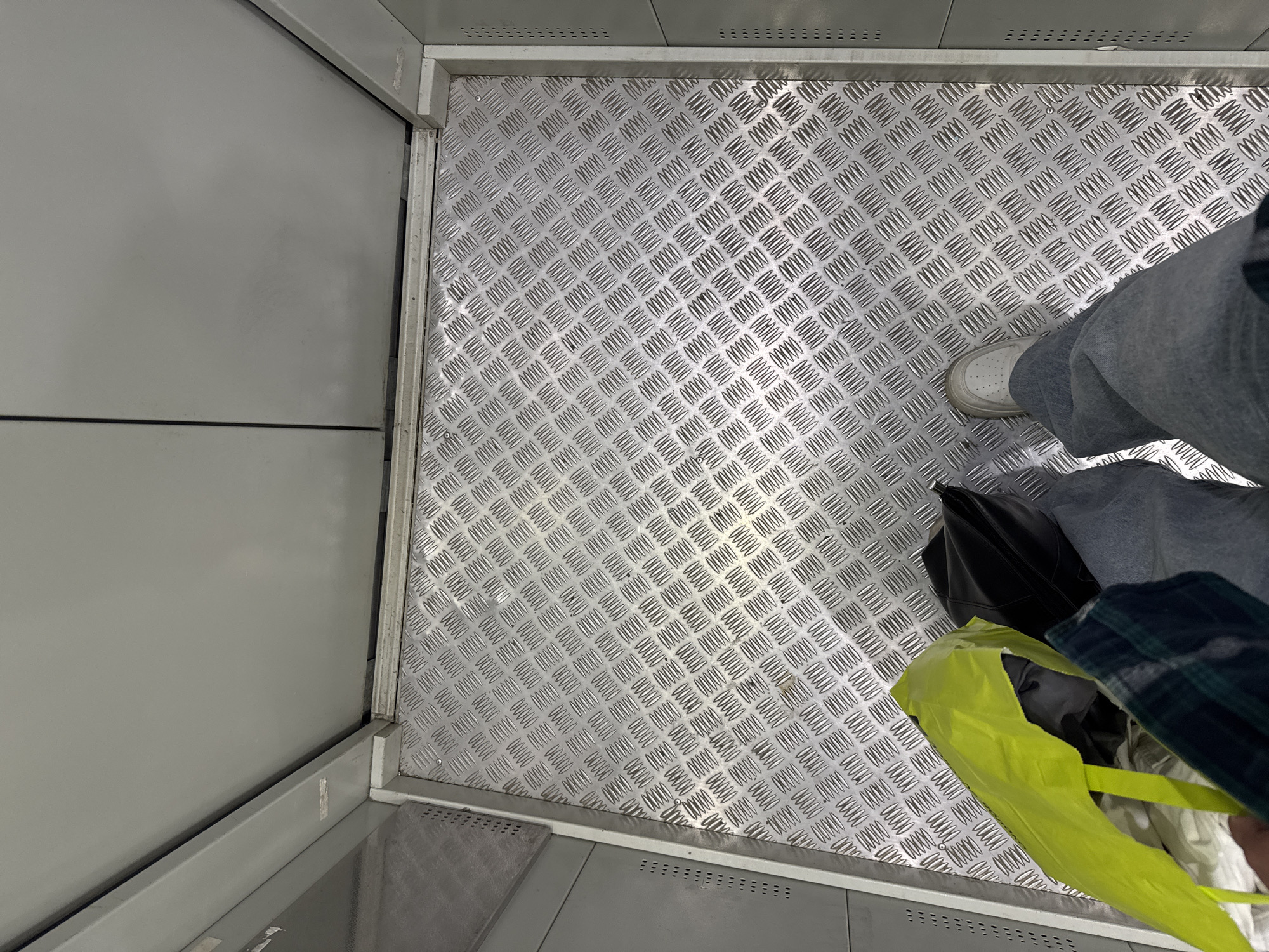 The image shows the interior of what appears to be an elevator or a similar confined space. The floor is covered with a diamond plate metal, providing a textured, industrial look. In the frame, we see the legs of at least two people, indicating they are standing in the space. One person is wearing grey pants and white sneakers. Another person's legs are visible, wearing grey pants and dark shoes, and they are holding a black bag. A bright yellow plastic bag is also present, partially obscuring some of the items. The walls of the space are a dull grey, with some visible paneling and what might be ventilation grilles at the bottom. The lighting suggests it is daytime, though it's difficult to determine the specific time of day due to the enclosed setting. The overall impression is utilitarian and functional, lacking any decorative elements. There is no discernible weather information or strong emotional cues visible. Some faint text, appearing as a series of numbers and symbols, is visible on the metal flooring, but it is too indistinct to be legible. The overall scene depicts people in a transit space, likely an elevator in a building.