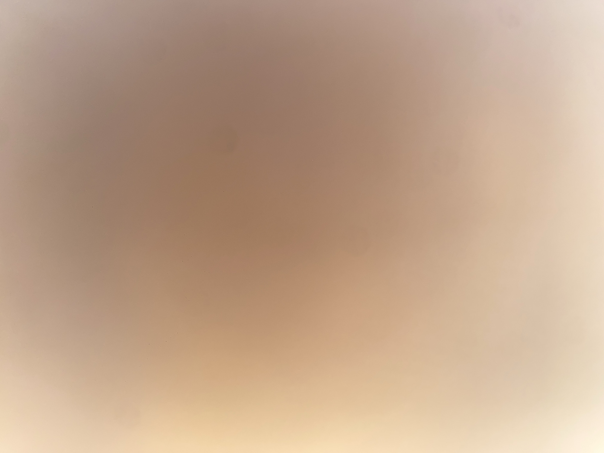 The image is a close-up, out-of-focus photograph of a surface. The surface is smooth and appears to be illuminated by a soft, warm light source, creating a gradient of color from a bright, pale yellow-orange on the left to a muted brown-orange on the right. There are several small, dark specks visible on the surface, likely dust or debris. The texture is uniform, suggesting a painted or fabric material. The image provides no discernible information about its location in Ablanitsa, Bulgaria, or any people, subjects, objects, or actions.