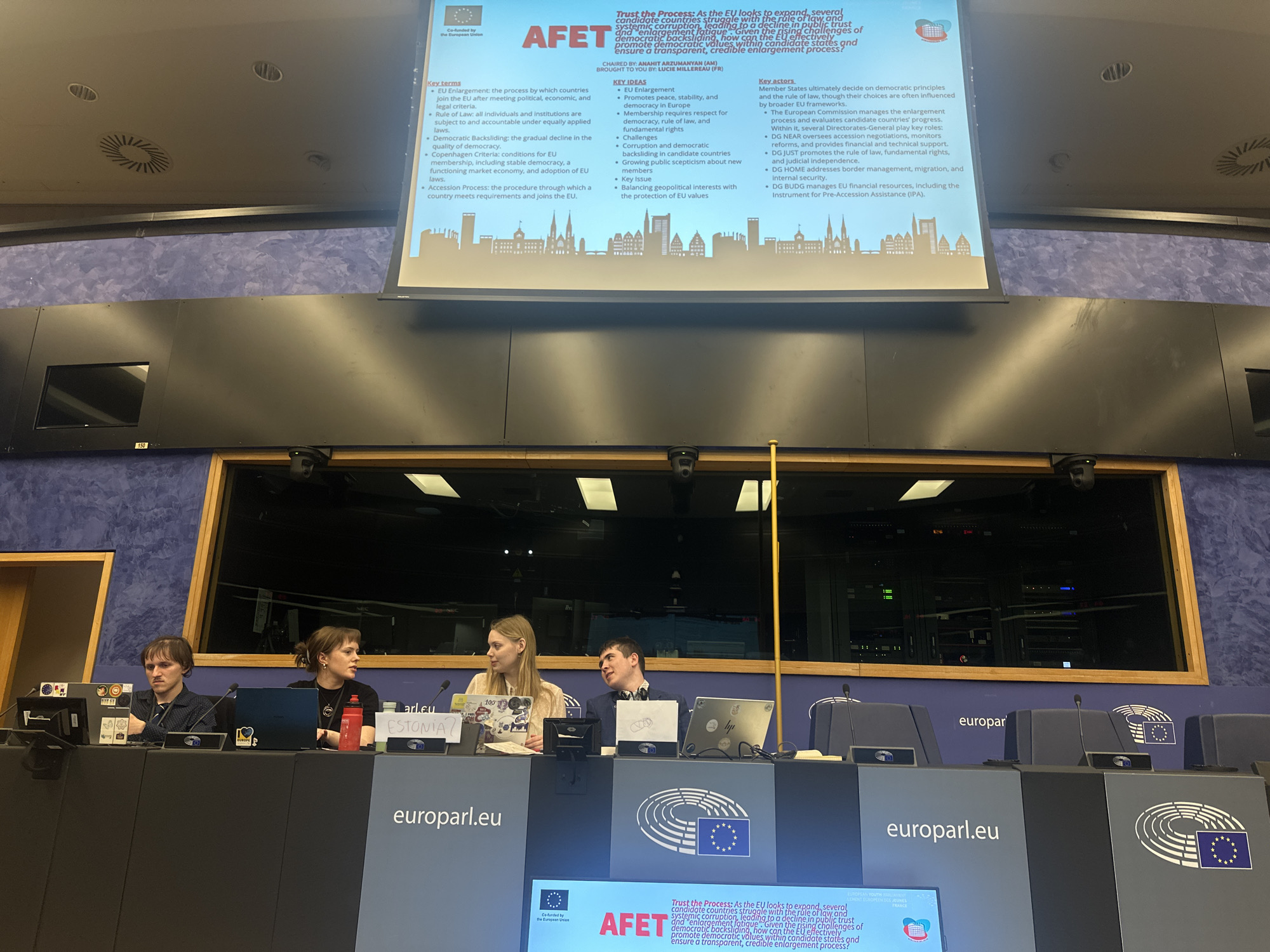 An indoor scene captures four young people, three women and one man, seated at desks arranged in a parliamentary or conference setting. They appear to be participants in an event, possibly a youth simulation or panel discussion, given the "europarl.eu" branding visible on the desks and a logo for "EUROPEAN YOUTH EVENT" on a small screen below the main display.
A large projector screen dominates the background, displaying a presentation slide titled "AFET" and "Trust the Process: As the EU looks to expand, several candidate countries struggle with the rule of law and systemic corruption, leading to a decline in public trust and enlargement fatigue. Given the rising challenges of democratic backsliding, how can the EU effectively promote democratic values within candidate states and ensure a transparent, credible enlargement process?" The slide is credited as "CHAIRED BY: ANAHIT ARZUMANYAN (AM)" and "BROUGHT TO YOU BY: LUCIE MILLEREAU (FR)." Key sections on the screen include "Key terms," "KEY IDEAS," and "Key actors," detailing concepts like EU Enlargement, Rule of Law, Democratic Backsliding, Copenhagen Criteria, Accession Process, and the roles of the European Commission and its Directorates-General (DG NEAR, DG JUST, DG HOME, DG BUDG). A "Co-funded by the European Union" logo is also visible.
The individuals are seated at individual desks equipped with microphones and laptops. Stickers on the laptops include "I love EUROPE" and an EU flag. One participant has a handwritten sign on their desk that reads "ESTONIA?". A red water bottle and a black one are also on the desks. The setting suggests a formal, educational, or political discussion, likely taking place in a European Parliament facility in Strasbourg, France.