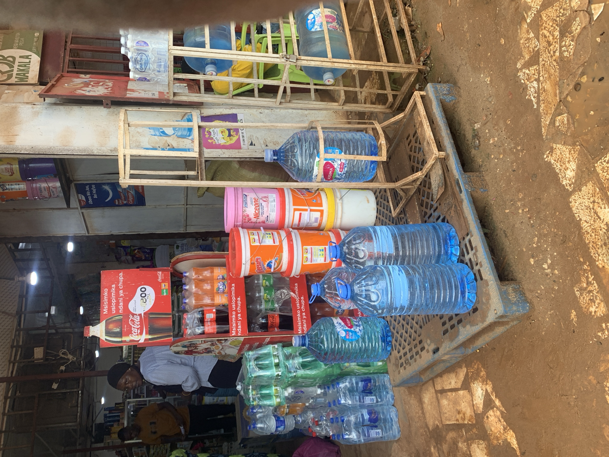 The image displays an outdoor retail setup in front of a small shop in Dar es-Salaam, Tanzania. The ground is unpaved, consisting of reddish-brown soil with embedded light-colored flagstones, showing patches of moisture and dirt.
In the immediate foreground, a blue plastic pallet supports several clear plastic bottles of water. Three large, multi-gallon blue-capped water bottles, one branded "KISIMA," are positioned prominently. Behind these, stacks of smaller individual bottled waters and multi-packs in clear plastic wrapping are visible.
To the left of the water display, a prominent red Coca-Cola point-of-sale display features a large Coca-Cola bottle image and the Swahili text "Msimsimo usiopimika ndani ya chupa." (Unquantifiable enjoyment inside the bottle.) and "Tsh 200". Below this, plastic bottles of Coca-Cola, Fanta, and Sprite are arranged in crates.
To the right, two light-colored, rudimentary wooden frame racks are present. One rack holds multiple large blue water bottles, some oriented horizontally, some branded "DESH MANIS." Another rack stands empty. Stacked near the Coca-Cola display and behind the water bottles are various colored plastic buckets, including orange "KLEESORS" and pink "Niceone" branded containers.
The background reveals the entrance to a small general store or duka. Inside, shelves hold assorted goods. Two individuals are present: a woman in a light-colored long-sleeved shirt and dark head covering stands with her back partially to the camera, looking left. A man in a brown t-shirt is partially visible deeper inside the shop on the left. Bare light bulbs illuminate the interior and overhang the entrance. Additional signage includes a blue sign with "Ubora Wa Juu, Bei Nafuu" (High Quality, Affordable Price) and a Colgate logo, a "WAKALA" sign on the upper right, and a "Ceres" juice poster on a pillar.
