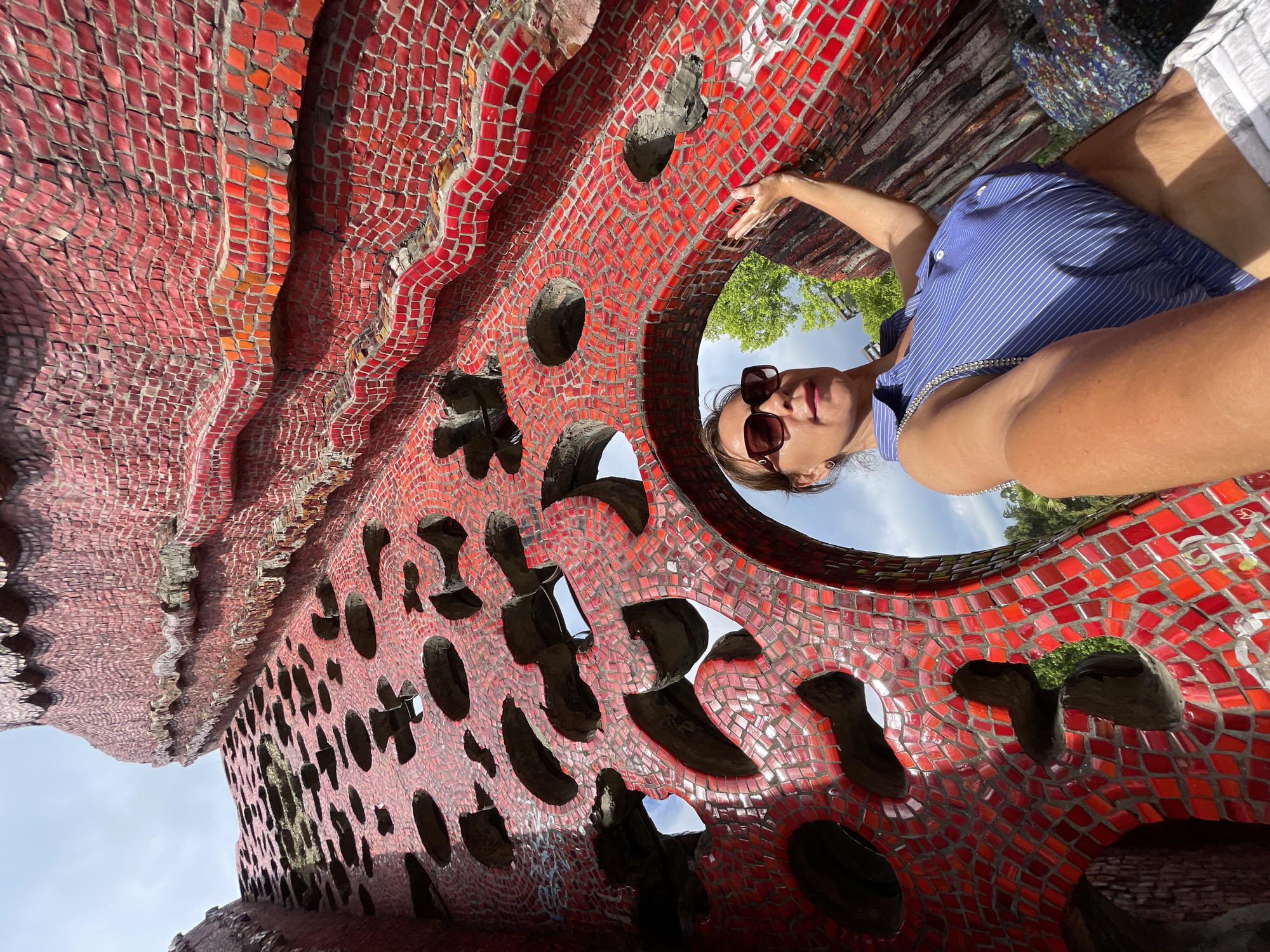 A woman takes a selfie in front of a vibrant red mosaic sculpture with organic, flowing shapes and numerous cutouts. The sculpture is made of small, irregularly shaped red tiles that create a textured, undulating surface. The woman is wearing a blue and white pinstripe sleeveless shirt and white shorts. She has on sunglasses and her right arm is extended, touching the sculpture. The background shows glimpses of green trees and a cloudy sky. This sculpture is located at the "Kukol'nyi Teatr Skazok" (Puppet Theater of Fairy Tales) in the city of Sochi, Russia, specifically in the central part of the city near the Arboretum.