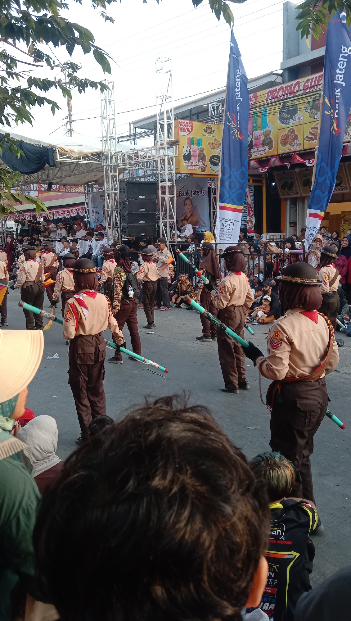 In the city of Jatisawit, Indonesia, a group of individuals, likely scouts given their uniforms and hats, are participating in an outdoor event. They are dressed in brown uniforms, including shirts, pants, and hats, and some are carrying long, cylindrical objects. The background reveals a street lined with buildings displaying banners and advertisements, including one for "Bank Jateng" and another with "PROMO HEBOH." A stage with sound equipment is visible to the left, and a crowd of spectators is gathered, watching the procession. The scene suggests a parade or a community gathering in a public space.