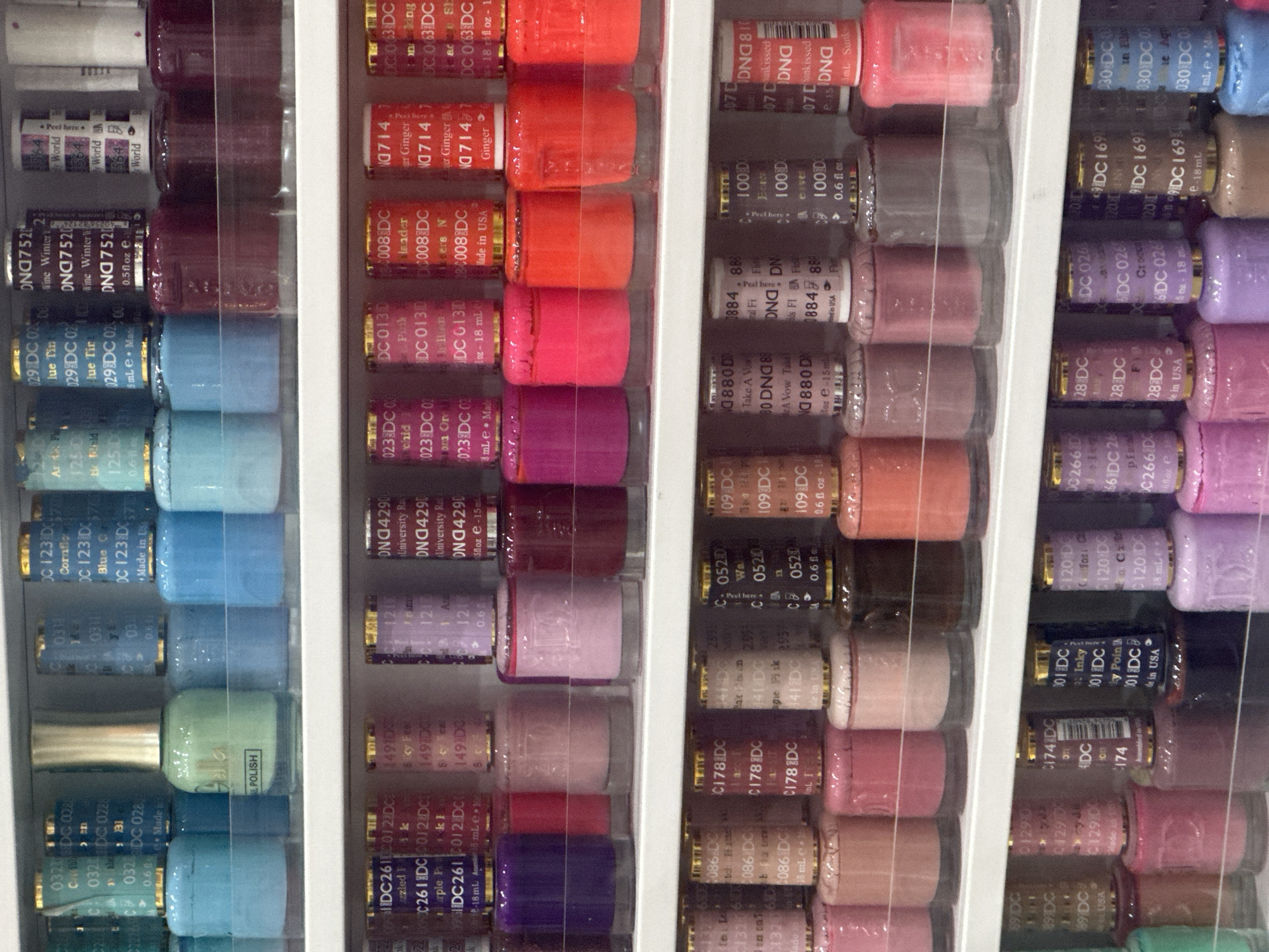 The image shows a display of nail polish bottles on white shelves. The bottles are of various colors, including green, blue, pink, purple, red, orange, and brown. Many of the bottles have labels with text and numbers, such as "DC 028", "DNA 7520", "DC 261", "DC 013", "DC 008", "DC 714", "DC 0520", "DC 109", "DC 880", "DC 100D", "DC 810", "DC 24", "DC 174", "DC 1290", "DC 026", "DC 169", and "DC 030". Some of the labels indicate "Made in USA" or "Made in China". The shelves are organized with multiple rows of bottles. The background is a white, out-of-focus surface. The image is a close-up of the nail polish display, and no people or any specific city or location information are visible within the frame. Therefore, it is not possible to determine if this image is located in Coffs Harbour, Australia.