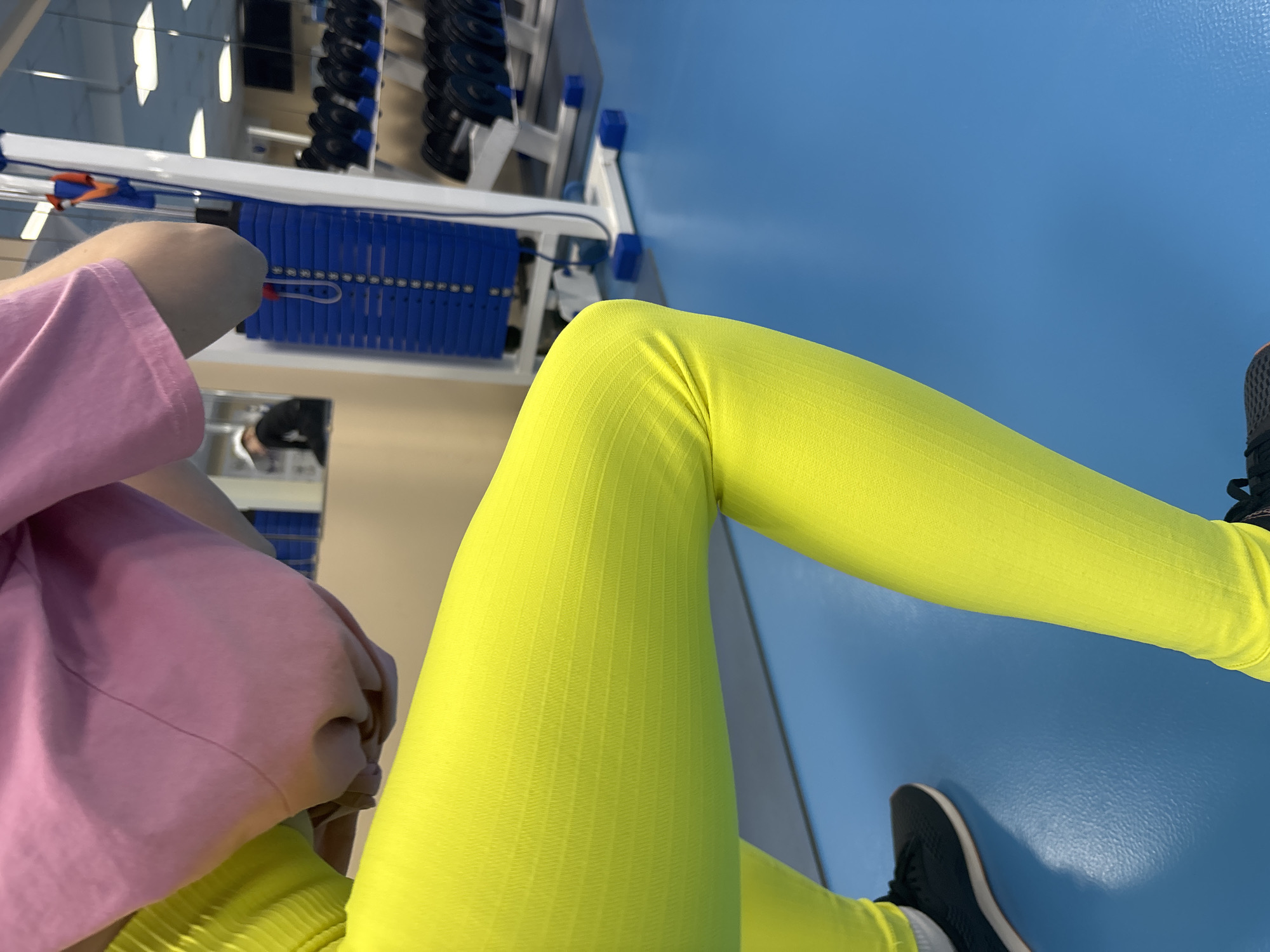 The image presents a view from above, featuring a person wearing bright yellow leggings and black sneakers. The person is wearing a pink and beige sweatshirt. They appear to be seated or lying down with one leg extended straight and the other bent at the knee. The setting seems to be a gym or fitness center in Chaikovsky, Russia, indicated by the blue flooring and weightlifting equipment visible in the background. The equipment includes a weight machine with blue weight plates and a rack with dumbbells.