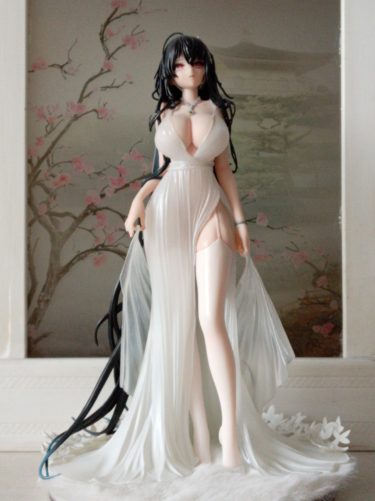 A full shot photograph shows a figurine of a woman standing against a backdrop featuring a painted scene of a tree with pink blossoms on the left and a mountainous landscape on the right. The figurine is that of a woman with long black hair, red eyes, and pale skin, wearing a sheer white dress with a plunging neckline. The dress has long sleeves and a thigh-high slit, revealing stockings and garters. She is adorned with a silver necklace. The figurine is standing on a white furry surface, with white floral accents at the base of the dress. The lighting is bright and diffused, casting minimal shadows. The image does not contain any information that would allow for its location to be identified in Yessentuki, Russia.