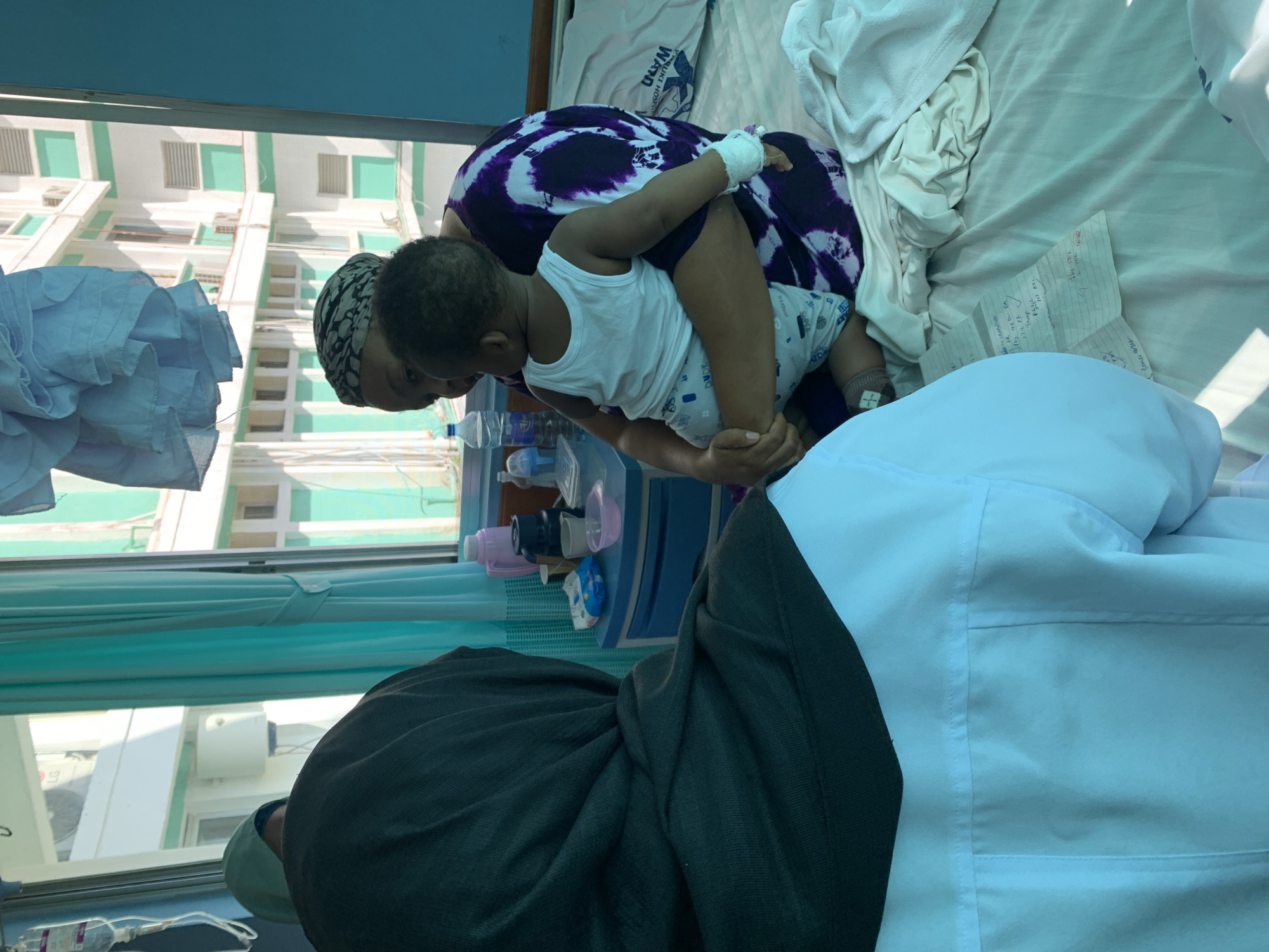 The image captures a scene within a hospital room, likely located in Dar es-Salaam, Tanzania. A woman wearing a headscarf and a purple and white patterned dress is holding a young child. The child wears a white tank top and patterned shorts. There is a medical IV line in the child's arm. The room contains a bed with white linens, medical equipment on a shelf near a window, and a person wearing a dark sweater and a light colored medical coat. The window reveals a building exterior painted in white and light green. The overall atmosphere suggests a hospital setting where a child is being cared for.