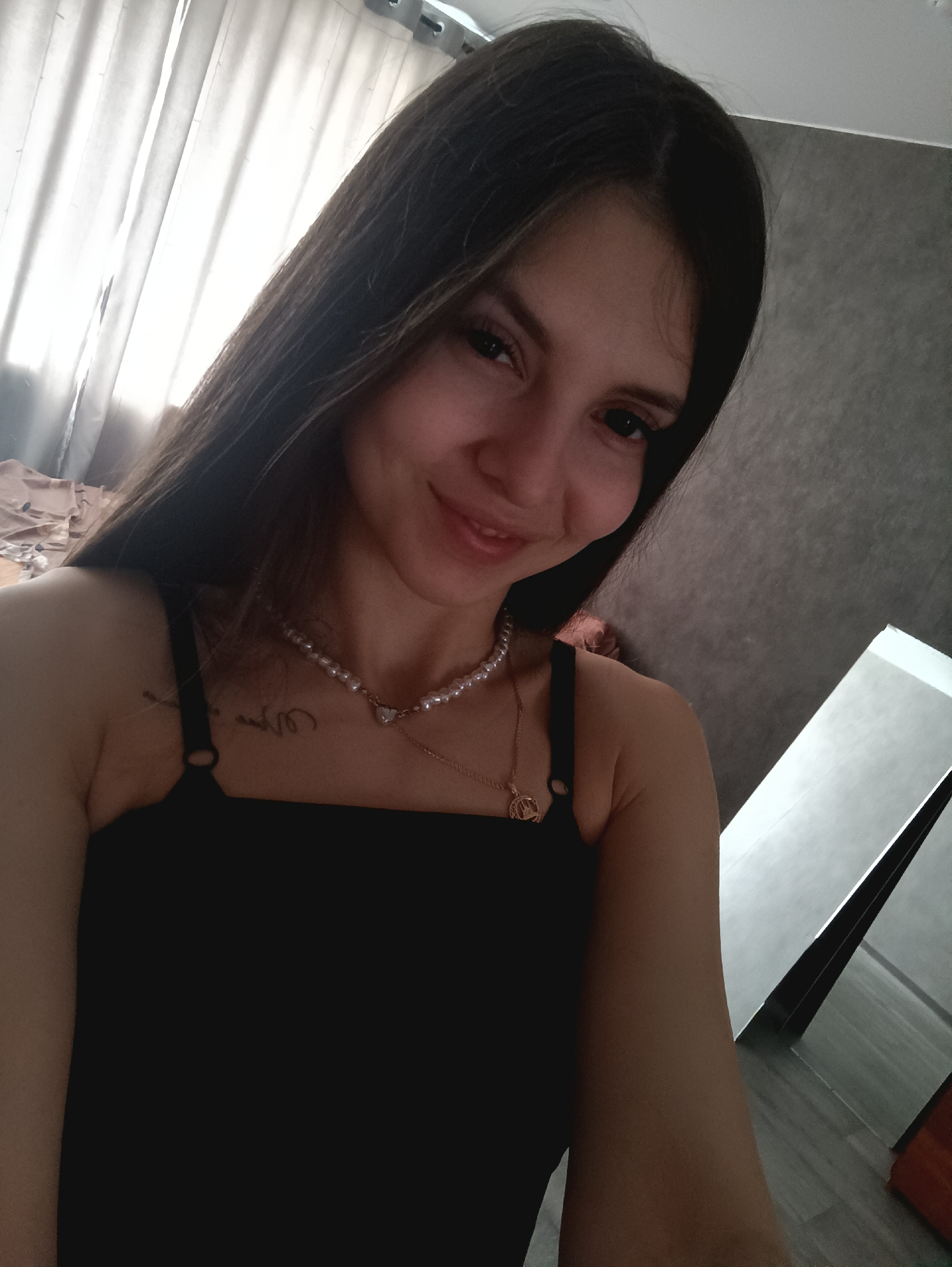 A woman with long brown hair is facing the camera and smiling. She is wearing a black top and a pearl necklace with a heart pendant, as well as a gold necklace with a circular pendant. She has a tattoo on her upper chest. In the background, there is a white curtain, a gray wall, and a white leaning object reflecting light. The location is inside an unspecified room.