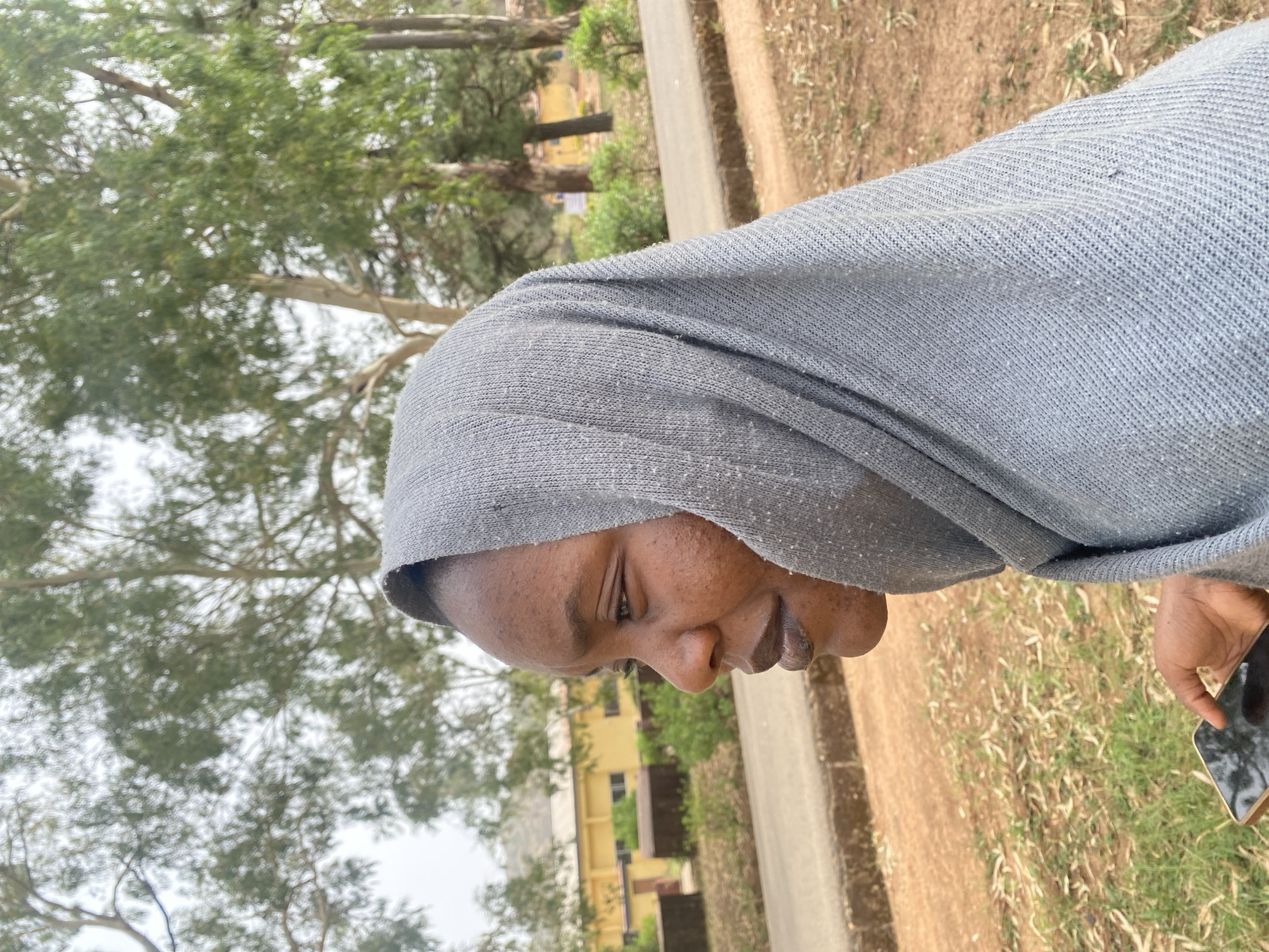 An outdoor, eye-level, portrait-oriented shot captures a person of color, possibly female, in profile, looking downwards. The individual is wearing a light gray or light blue head covering that also drapes over the shoulders. Their dark skin is visible, with some texture on the forehead and cheek. In the lower right corner, their right hand holds a black smartphone with a reflective screen.
The background features lush green trees with visible branches and leaves, suggesting a natural or campus environment. Behind the person and the trees, a light yellow building with dark windows is partially visible in the mid-ground. To the right, a dirt path or road runs alongside a patch of grassy, somewhat dry ground. The lighting is soft and diffused, indicating an overcast day. No specific activity beyond holding a phone is discernible. There is no visible text. The scene is set in Bukuru, Nigeria.