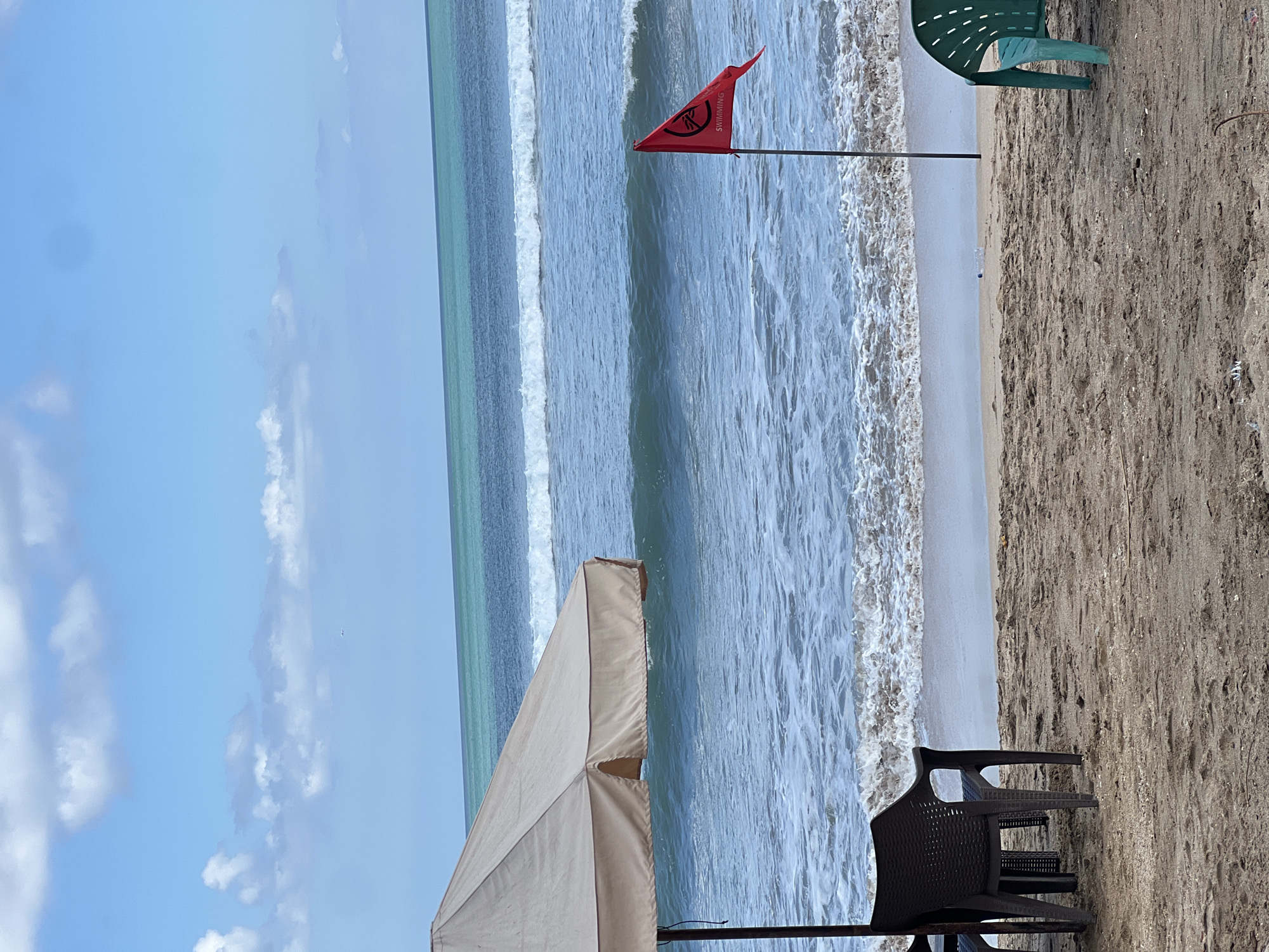 The image depicts a beach scene in Kuta, Indonesia, with the ocean and sky dominating the frame. In the foreground, there is a beige umbrella providing shade over two dark plastic chairs. To the right, a red flag with a symbol and the word "SWIMMING" is planted in the sand, indicating a warning. A green plastic chair is visible further to the right. The ocean displays moderate waves breaking on the shore, with a turquoise hue in the distance. The sky is blue with scattered white clouds, and a distant airplane can be seen. The sandy beach is relatively empty, with no people visible in the immediate vicinity. The overall atmosphere suggests a potentially hazardous swimming condition due to the red flag.