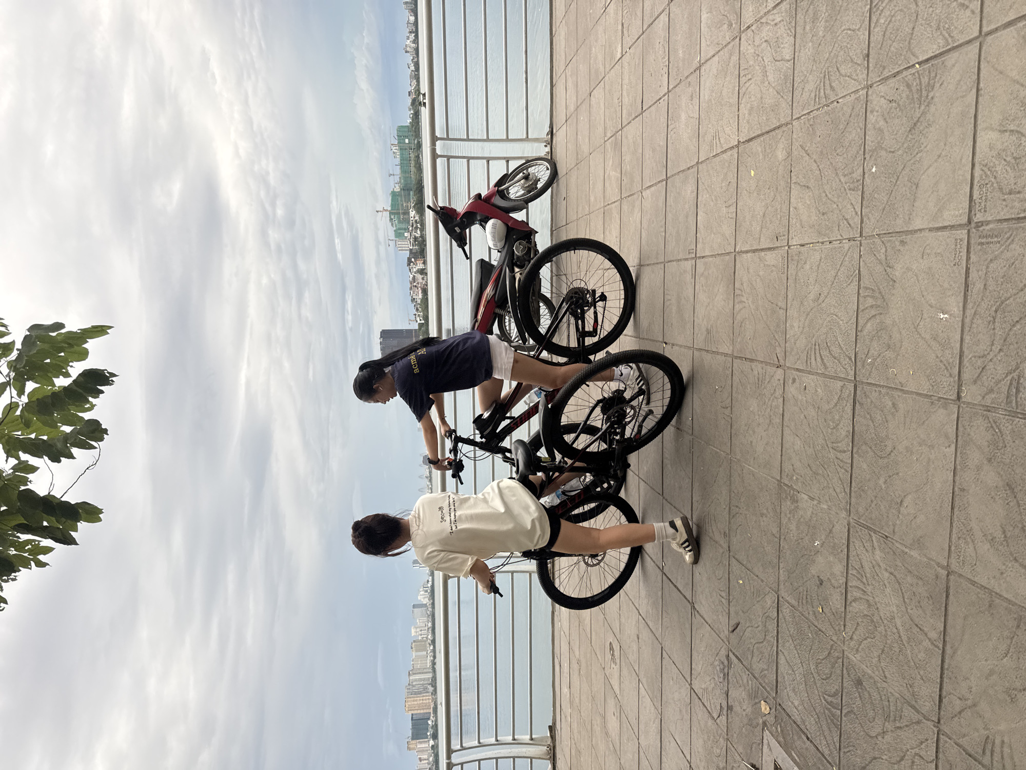 The image depicts two young women by a waterfront promenade in Hanoi, Vietnam. The woman in the foreground, facing away from the camera, wears a cream-colored t-shirt with text and black shorts. She stands next to a black bicycle, her left hand on the handlebars. The second woman, also facing away, is positioned to the right of the first. She wears a dark t-shirt with text and white shorts, and is also near a bicycle. A red and silver motorcycle is parked behind the two women. The promenade is paved with textured square tiles. Beyond the railing, a body of water is visible, with a cityscape featuring buildings and construction cranes in the distance. The sky is overcast.