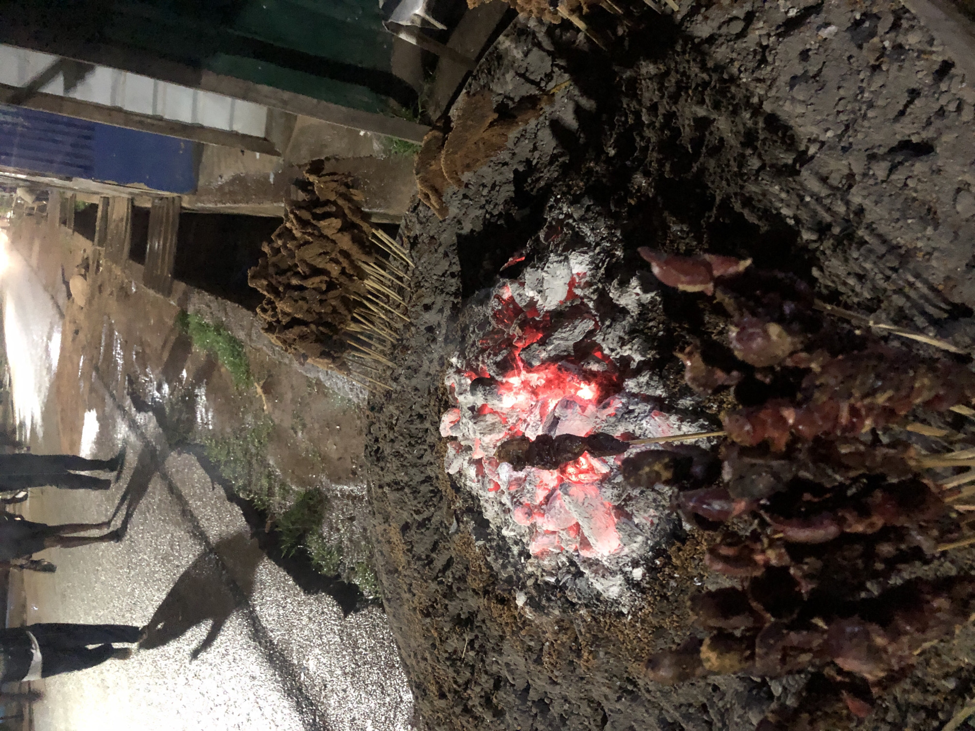 This nighttime image captures a street food vendor's setup in Unknown, Unknown. In the foreground, a large mound of dark earth or ash serves as a base for grilling, with a central cluster of glowing red-orange hot coals. Numerous wooden skewers, laden with dark brown to reddish meat or offal, are arranged around the coals, with a denser group visible in the lower left. To the right of the cooking area, a drainage ditch runs parallel to a wet, reflective asphalt road. In the background, three individuals walk away from the viewer on the road, casting elongated shadows. Portions of a building with a blue section are visible on the far right. Sparse green grass can be seen near the ditch. The scene is illuminated by artificial light sources, creating strong reflections on the wet ground.