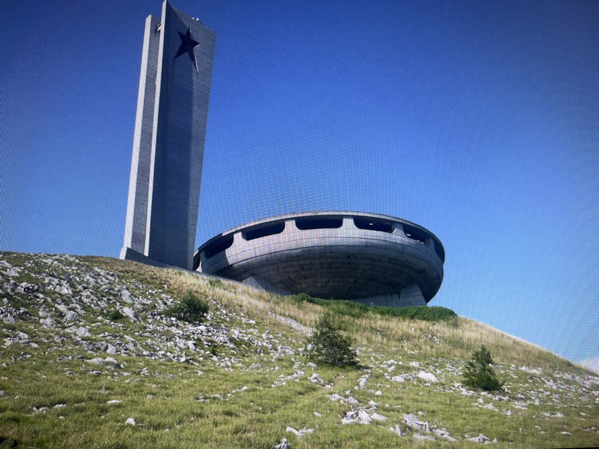 The image showcases a monumental, brutalist architectural complex set on a steep, rocky hillside under a clear, bright blue sky, characteristic of a sunny day in Varna, Bulgaria. The scene is devoid of people, emphasizing the imposing scale and desolate surroundings of the structures.
The complex consists of two primary elements. To the left, a tall, slender rectangular pylon or tower rises dramatically, tapering slightly towards its apex. A large, dark five-pointed star emblem is prominently affixed to one of its upper sides, suggesting a historical or ideological significance often associated with Soviet-era architecture. The pylon itself appears to be constructed from grey concrete with subtle vertical indentations or panels.
Adjacent to the pylon and further down the slope, a massive, circular, saucer-shaped building is partially embedded into the hillside. This striking structure, also seemingly made of concrete, features a continuous band of dark, elongated window-like openings or recesses around its upper circumference. Its futuristic and somewhat derelict appearance hints at a past grand purpose now left to the elements.
The natural setting comprises a rugged, arid hillside covered with sparse patches of green and dry grass, heavily interspersed with numerous grey and white rocks and boulders of varying sizes. A few small, scattered green bushes or stunted trees dot the landscape, adding to the sense of isolation and raw nature surrounding the man-made marvel. The overall impression is one of stark grandeur and a testament to a bygone era. No visible text is present in the image.