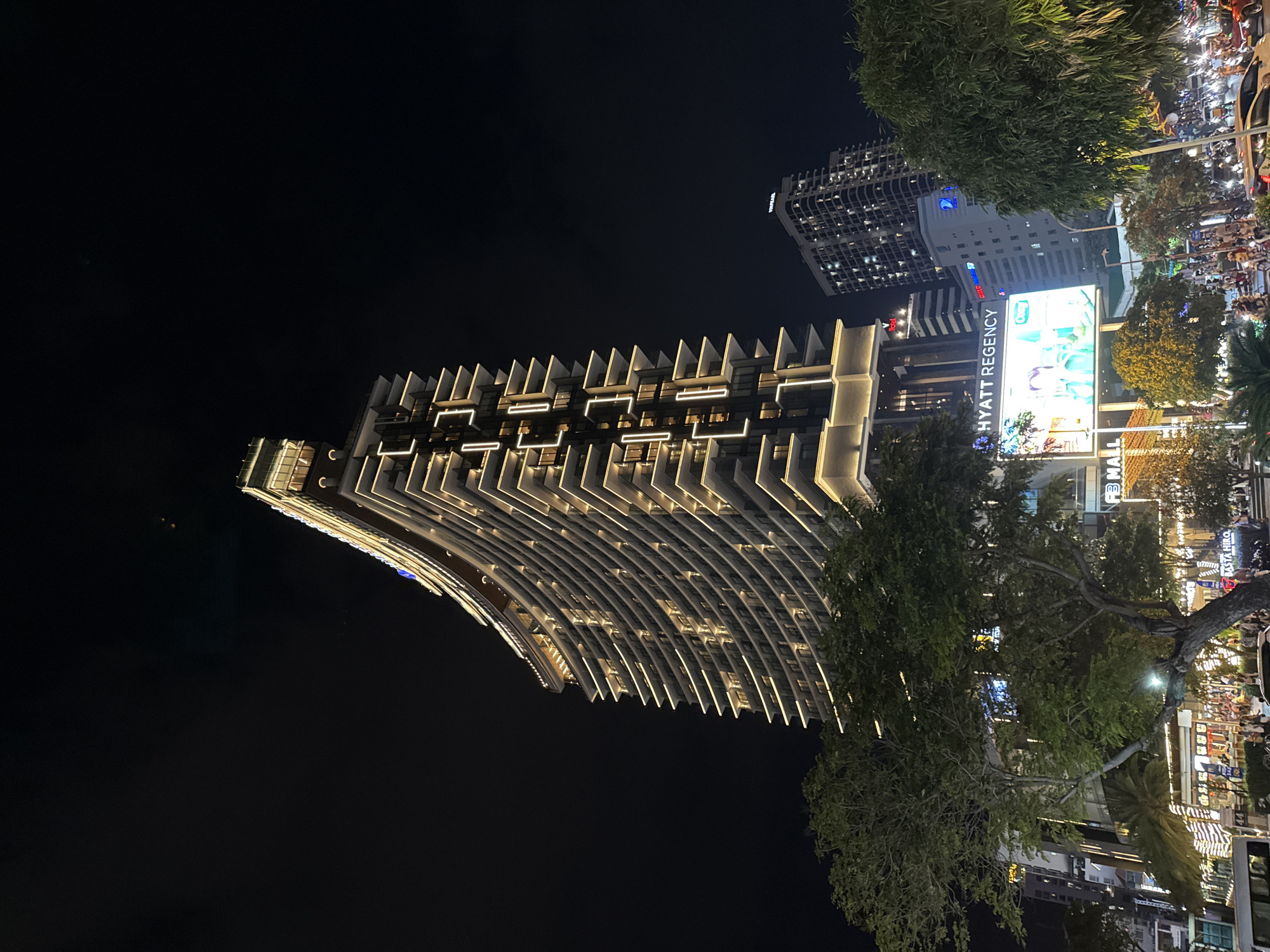 This image captures a nighttime view of the Hyatt Regency hotel in Nha Trang, Vietnam. The main subject is the distinctive curved architecture of the hotel, illuminated by warm exterior lighting that highlights its numerous balconies and vertical decorative elements. In the foreground, lush green trees partially obscure the lower levels of the hotel and the street scene below.
The street in front of the hotel is bustling with activity. Motorbikes and cars are visible, suggesting significant traffic flow, and a large digital billboard advertising "Hyatt Regency" is prominently displayed. Other buildings, including a taller, darker high-rise and a lighter-colored building with many windows, are visible in the background. The overall atmosphere is one of a vibrant urban environment at night.