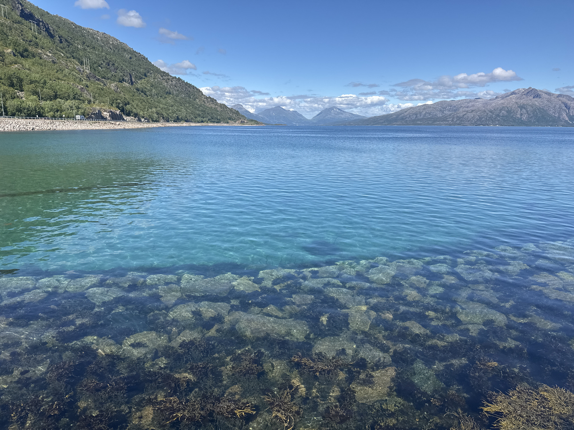 This media file depicts a scenic seascape in Lødingen, Norway, under a clear blue sky with scattered white clouds. The foreground features shallow, crystal-clear turquoise water revealing a rocky seabed with visible seaweed. The water transitions to a deeper blue further out.
The middle ground is dominated by a wide expanse of calm, blue water. In the background, a chain of majestic mountains, some with snow-capped peaks, stretches across the horizon. A road with a white campervan is visible running along the base of a green, tree-covered mountain to the left of the frame. This mountain is dotted with utility poles and wires. The overall scene evokes a sense of tranquility and natural beauty.
The time of day appears to be midday, given the bright sunlight and clear sky. The weather is fair and pleasant. The presence of mountains and the distinct coastline strongly suggest a Norwegian fjord or coastal landscape. No text is visible in the image.