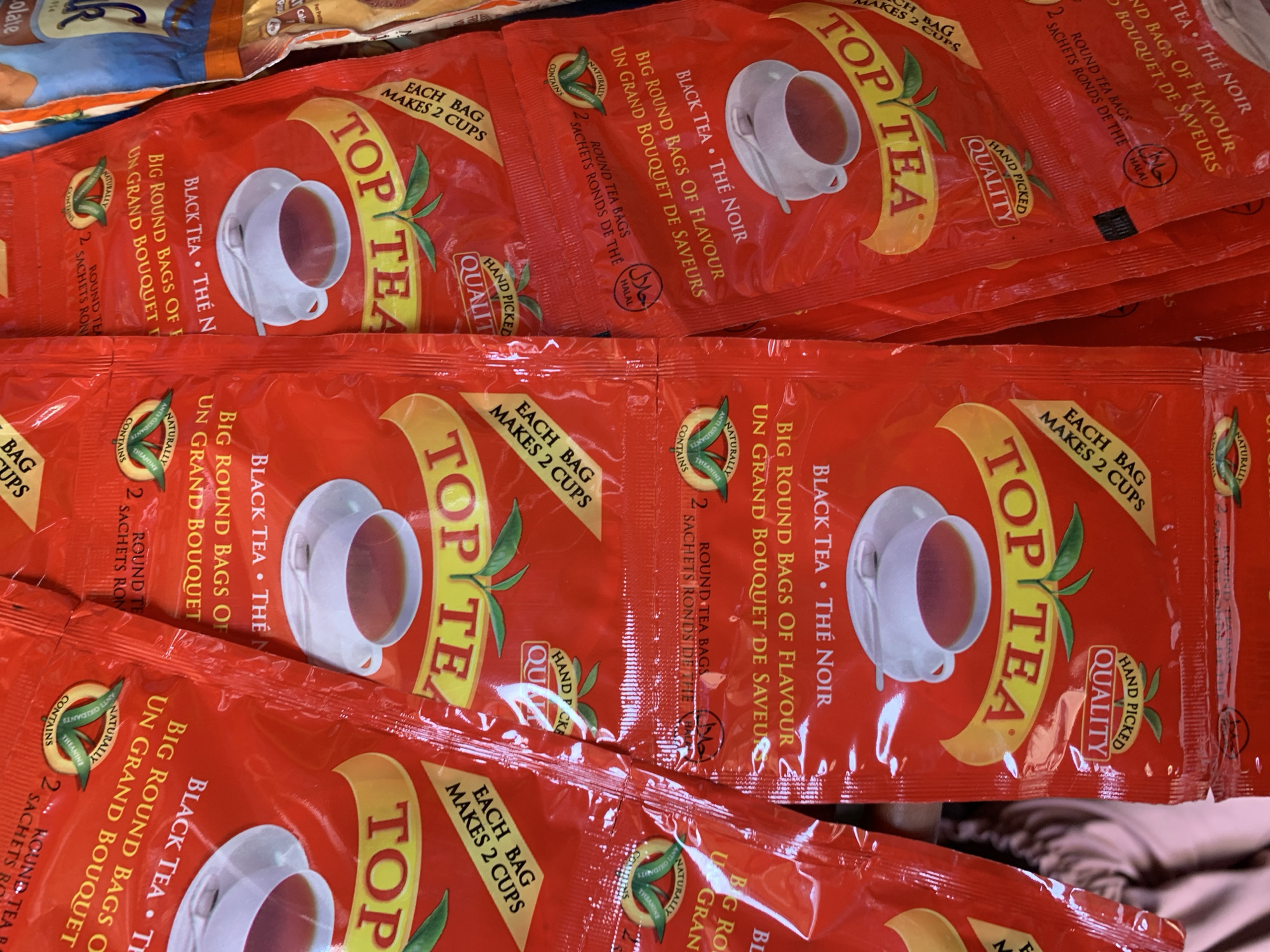 The image shows several packets of "Top Tea" brand black tea. Each red packet features a white teacup with tea, and text in English and French indicating it's "Black Tea - Thé Noir" and "Big Round Bags of Flavour - Un Grand Bouquet de Saveur." Each bag makes 2 cups, contains theanine, and is halal certified. They are packaged as 2 round tea bags per individual packet. The location, Rafin Par, Nigeria, is not discernible from the image.