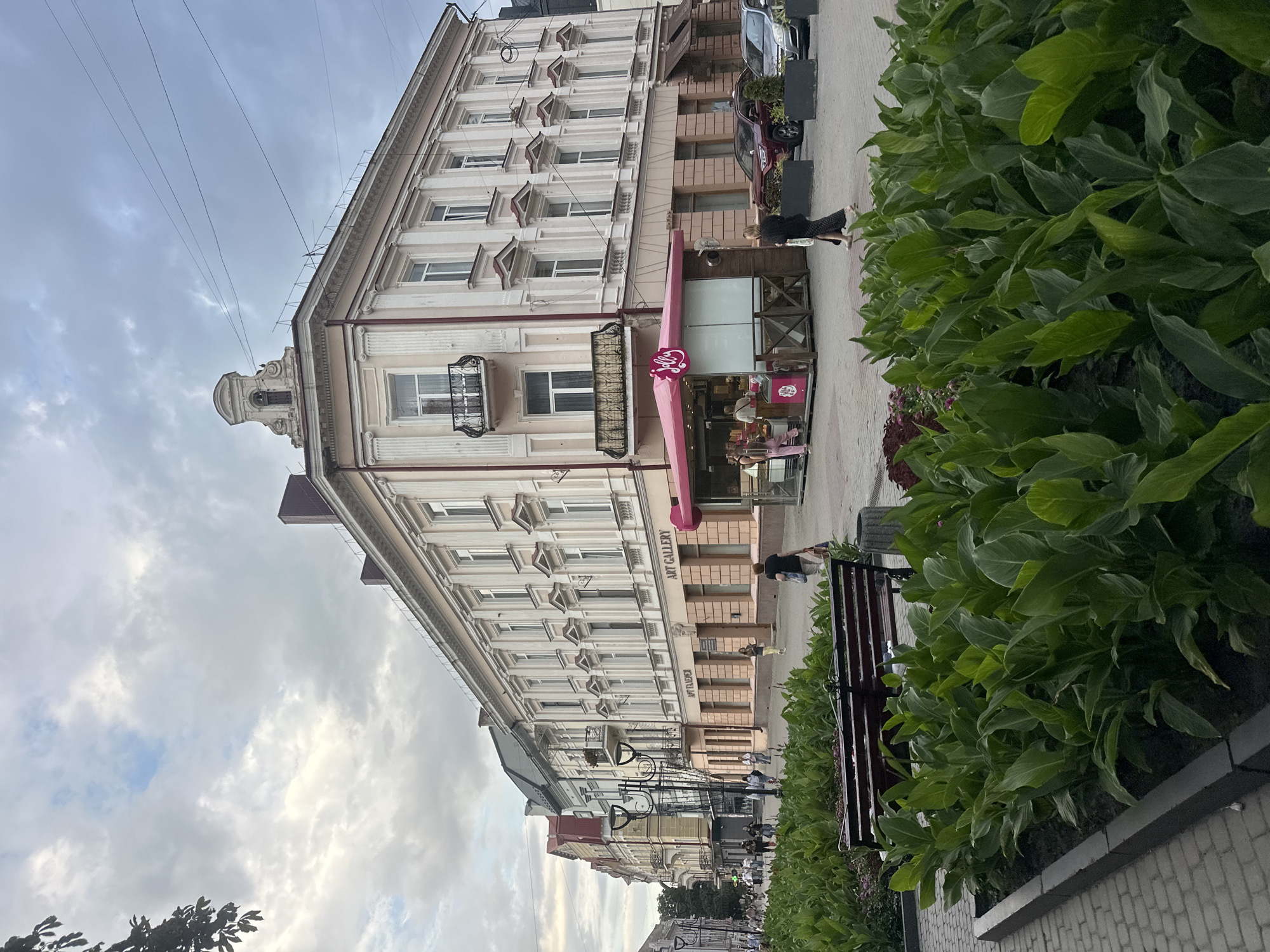 The image depicts a street scene in Ternopil, Ukraine, with a focus on the architecture and pedestrian activity. In the foreground, a well-maintained planter box filled with lush green plants is prominent, with a dark wooden bench to its left. Beyond the greenery, a paved pedestrian walkway stretches into the distance. On the right side of the walkway, a small pink kiosk with the logo "Jelly" is visible, suggesting it is a food or beverage vendor. Next to the kiosk, several vehicles are parked, including a dark red SUV and a silver sedan.
Dominating the mid-ground and background is a grand, multi-story building with a light-colored facade, adorned with architectural details like columns, ornate window frames, and a distinctive decorative element at the top. The building features numerous windows, some with balconies. Signs on the lower level indicate businesses, including one with "ART GALLERY" in English and Ukrainian lettering "АPT ГAЛЕРЕЯ". A line of other buildings, varying in architectural style and color, extends down the street to the left, creating a dense urban environment.
Several individuals are present in the scene. A woman in dark clothing walks from right to left in front of the kiosk, carrying a shopping bag. Another woman in pink pants is visible behind the counter of the kiosk, interacting with customers. Other pedestrians are seen walking along the street in the distance. The sky is overcast with a mix of grey and lighter clouds, and power lines are visible overhead. The overall atmosphere is that of a typical European city street with a blend of commercial activity and public space.