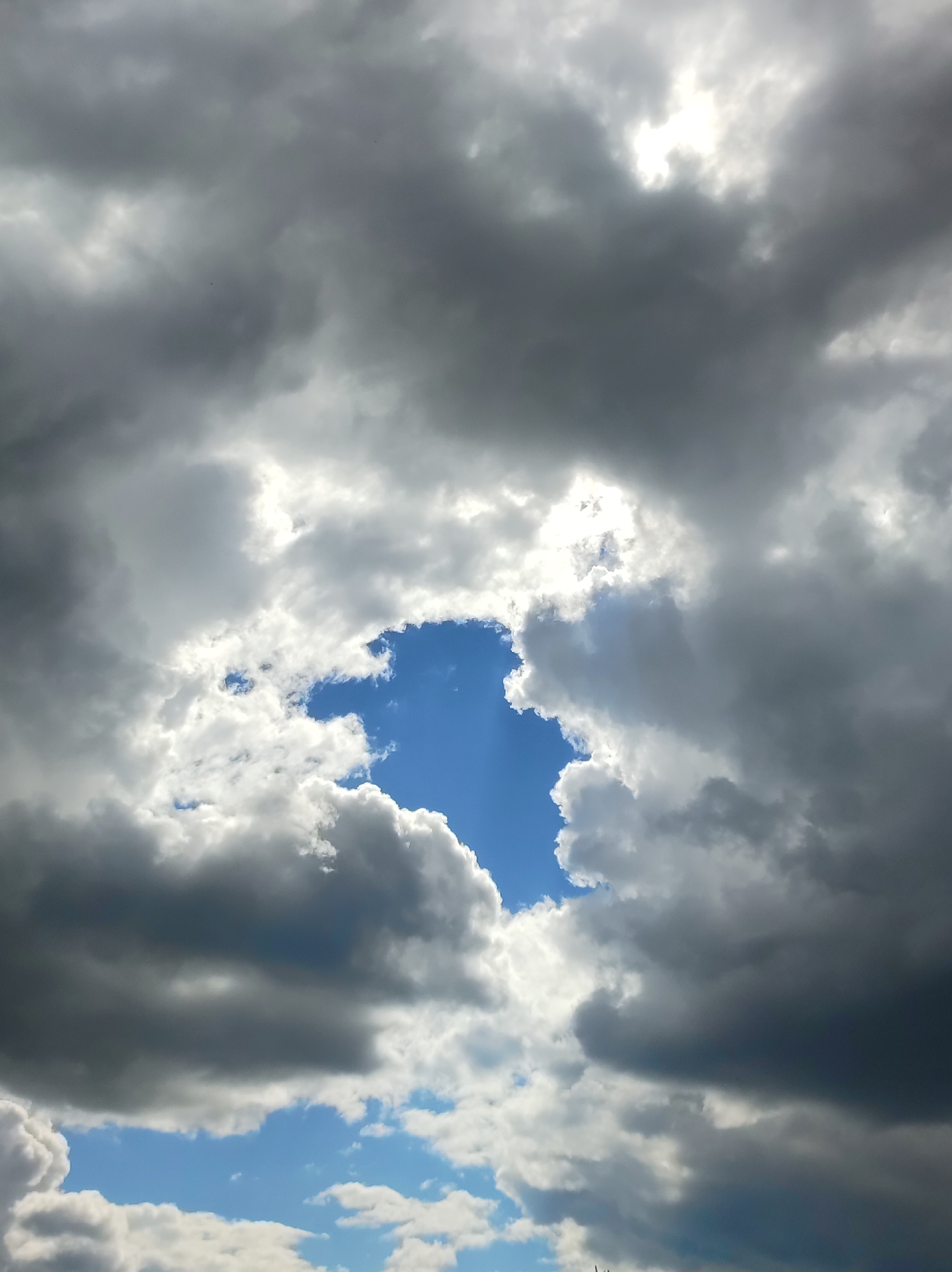The image is a photograph of the sky, with a significant portion covered by dark, heavy clouds. In the center of the frame, there is an opening in the clouds that reveals a bright blue sky. Lighter, fluffier clouds are visible around the edges of this opening, and some sunlight is breaking through in the upper right portion of the image. The lower portion of the image shows patches of blue sky with scattered white clouds. The scene is of a natural environment, specifically the sky above the city of Fokino, Russia. There are no visible people, subjects, objects, actions, or interactions depicted in the photograph.