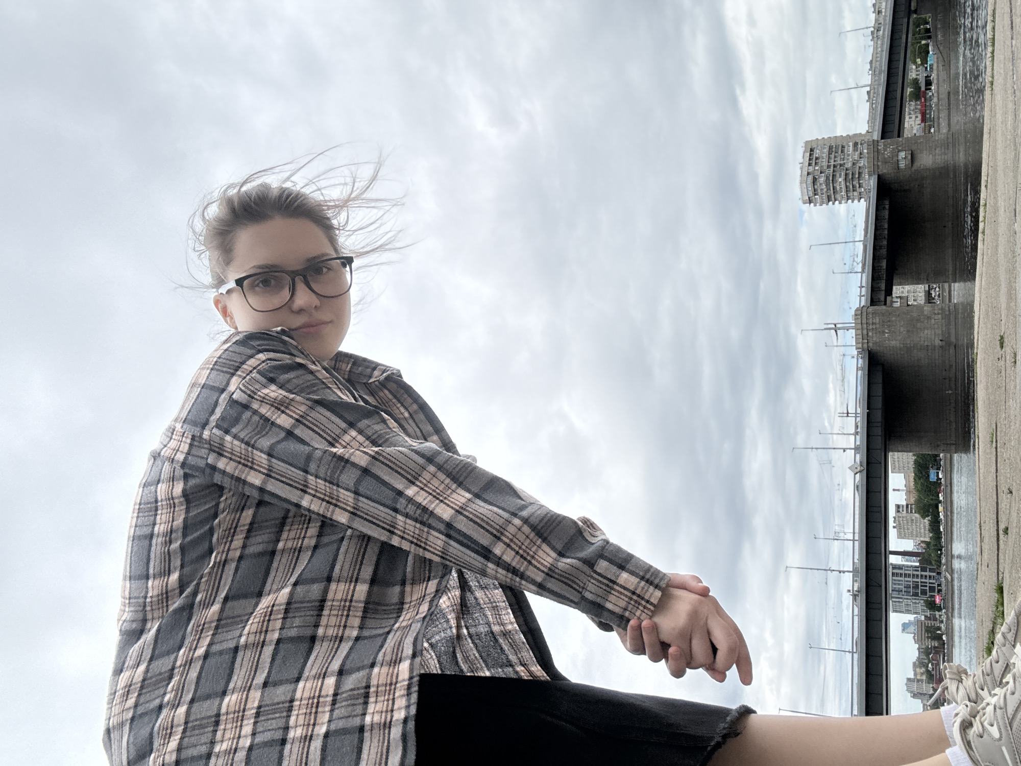 A person is captured in a low-angle selfie, looking down at the camera. They are wearing a plaid shirt and glasses. Their hands are clasped in front of them. In the background, a bridge spans a body of water, with city buildings and a distinctive apartment building visible on the other side. The sky is overcast.