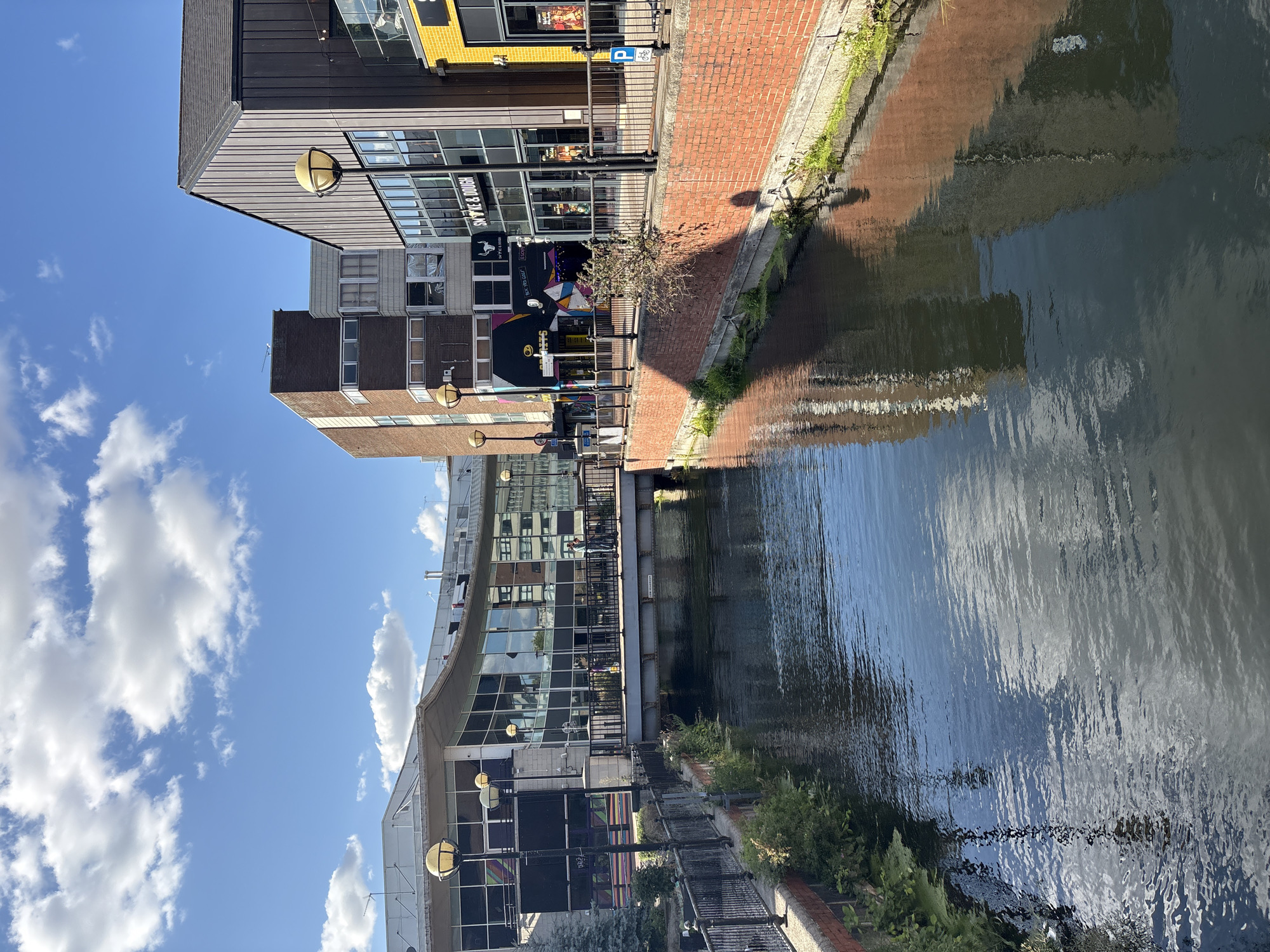 The image shows a view of the River Chelmer in Chelmsford, United Kingdom, looking towards the city center. The river flows through a channel lined with brick walls and bordered by a walkway with black metal railings. On the right bank, there are modern buildings with large windows, including a brick-faced building and a building with a dark metal facade. One of the buildings features a sign for "SMOKE & DOUGH". A street lamp with a spherical yellow light is visible on the walkway. On the left bank, there is a contemporary building with extensive glass windows, reflecting the sky and surrounding structures. A pedestrian bridge spans the river in the mid-ground. Several people are visible on the bridge, and some are walking along the walkways. The sky is blue with scattered white clouds, and the water reflects the sky and buildings, creating a sense of depth and urban scenery. Lush green vegetation grows along the edges of the river.