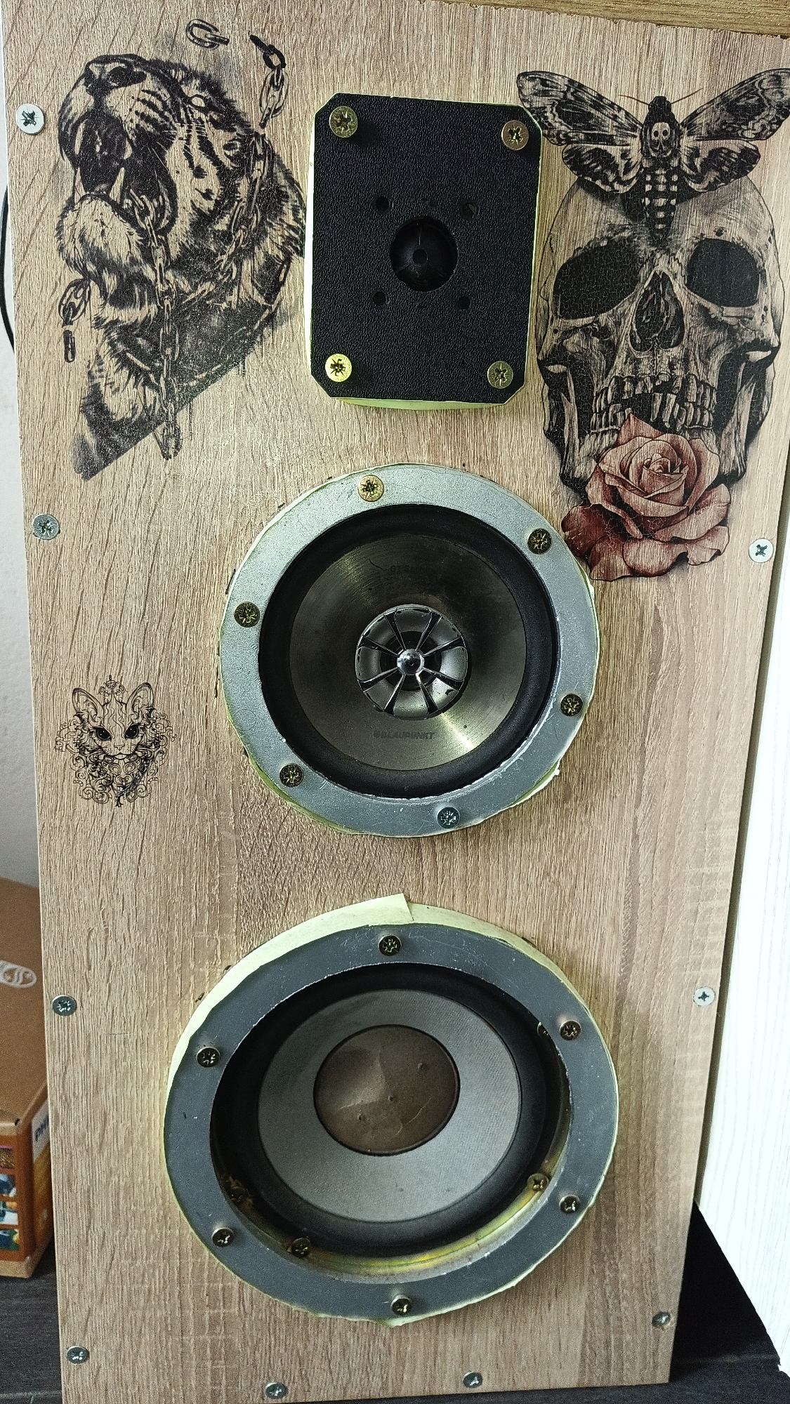 The image shows a close-up of a speaker cabinet. The cabinet is made of light-brown wood and has a grainy texture. Two circular speakers of different sizes are visible, each mounted with screws and surrounded by a silver frame with some tape around the bottom speaker. Above the larger speaker is a tweeter. The wood surface is decorated with various black and white graphics, including a tiger in chains, a skull with a moth on its head and a rose in its mouth, and a small stylized cat face. The image was not taken in Karnobat, Bulgaria.