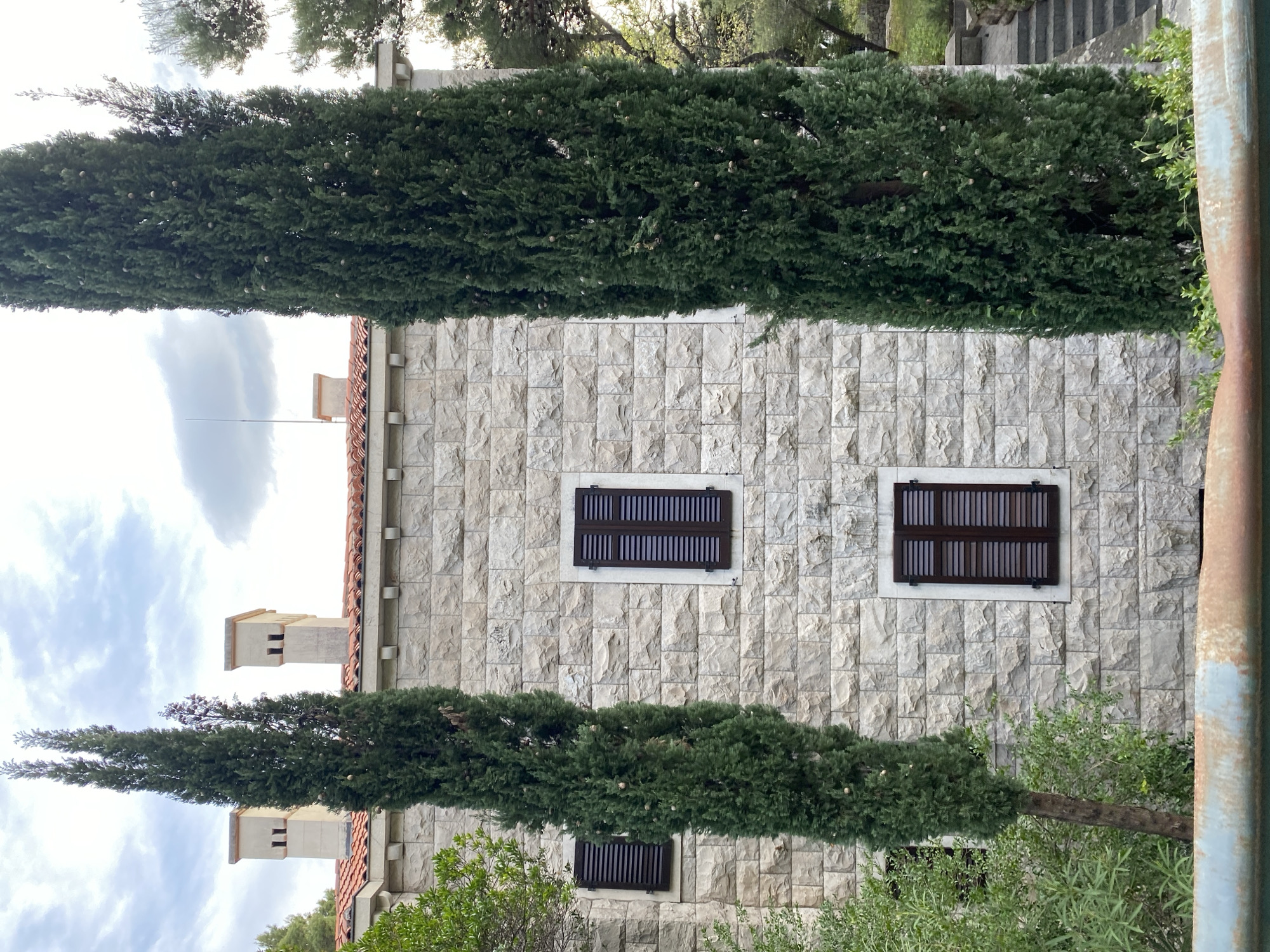 Exterior shot of a building with a stone facade, featuring two visible stories. The building has at least two rectangular windows with dark brown, louvered shutters. Tall, green, cypress-like trees flank the building on either side. A red-tiled roof is visible, with chimneys on top. The sky is cloudy. The location is Sveti Stefan, Montenegro, based on the architectural style and vegetation.