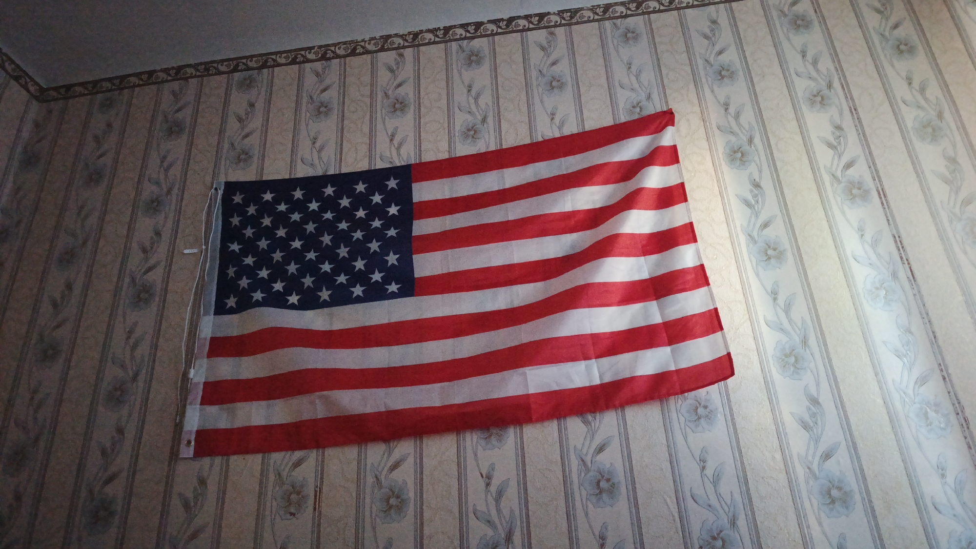 An American flag is draped on a wall. The flag is partially unfurled, showing its red and white stripes and the blue field with white stars. The wall behind the flag has a floral patterned wallpaper. The lighting in the image suggests an indoor setting, possibly a room. The location in the city of Barysh, Russia is not discernible from the visual information provided in the image.