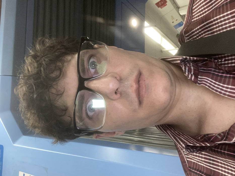 An adult male is depicted in a close-up, frontal self-portrait within a public transport vehicle in Luis Guillón, Argentina. The subject has dark, curly hair and is wearing black-framed glasses, through which his wide-open eyes with visible pupils are staring directly forward. Bright, rectangular light reflections are prominent in both lenses of his glasses. He is dressed in a collared, button-up shirt with maroon and white vertical stripes. A dark strap, consistent with a bag, is visible over his right shoulder. The environment suggests the interior of a train or subway car, featuring a light blue wall panel on the left with a partially visible white sticker, and a light-colored ceiling equipped with linear and circular overhead light fixtures. Dark horizontal structural elements are partially visible in the upper right background. No specific external landmarks or station identifiers are visible.