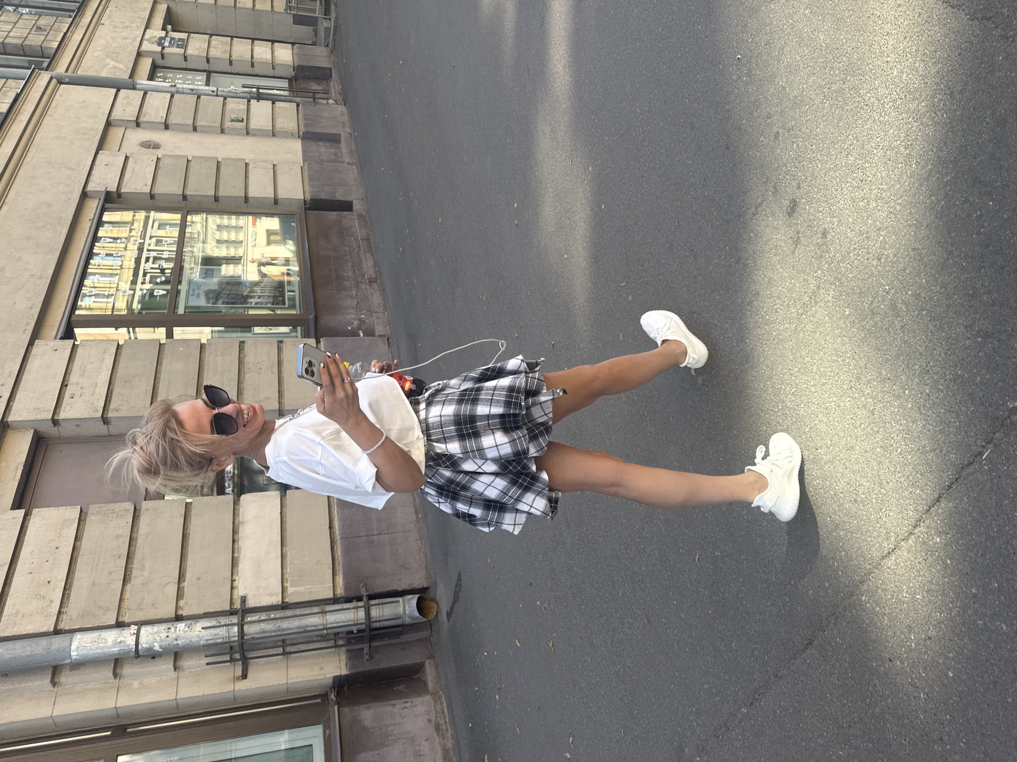 A woman with blonde hair styled in a messy bun, wearing large dark sunglasses, a white t-shirt, and a black and white checkered skirt, is walking on a paved city street. She is looking at her phone, which she holds in her right hand, and has a bottle with a red label in her left hand. She is wearing white sneakers. A white cable hangs from her phone. The scene appears to be in Saint Petersburg, Russia, as evidenced by the architectural style of the buildings in the background, characterized by classical detailing and large windows with reflections of other buildings. The buildings are light-colored and appear to be made of stone. A downspout runs vertically down the side of one of the buildings. The street is asphalt and appears to be well-maintained, with some shadows from sunlight falling on it.